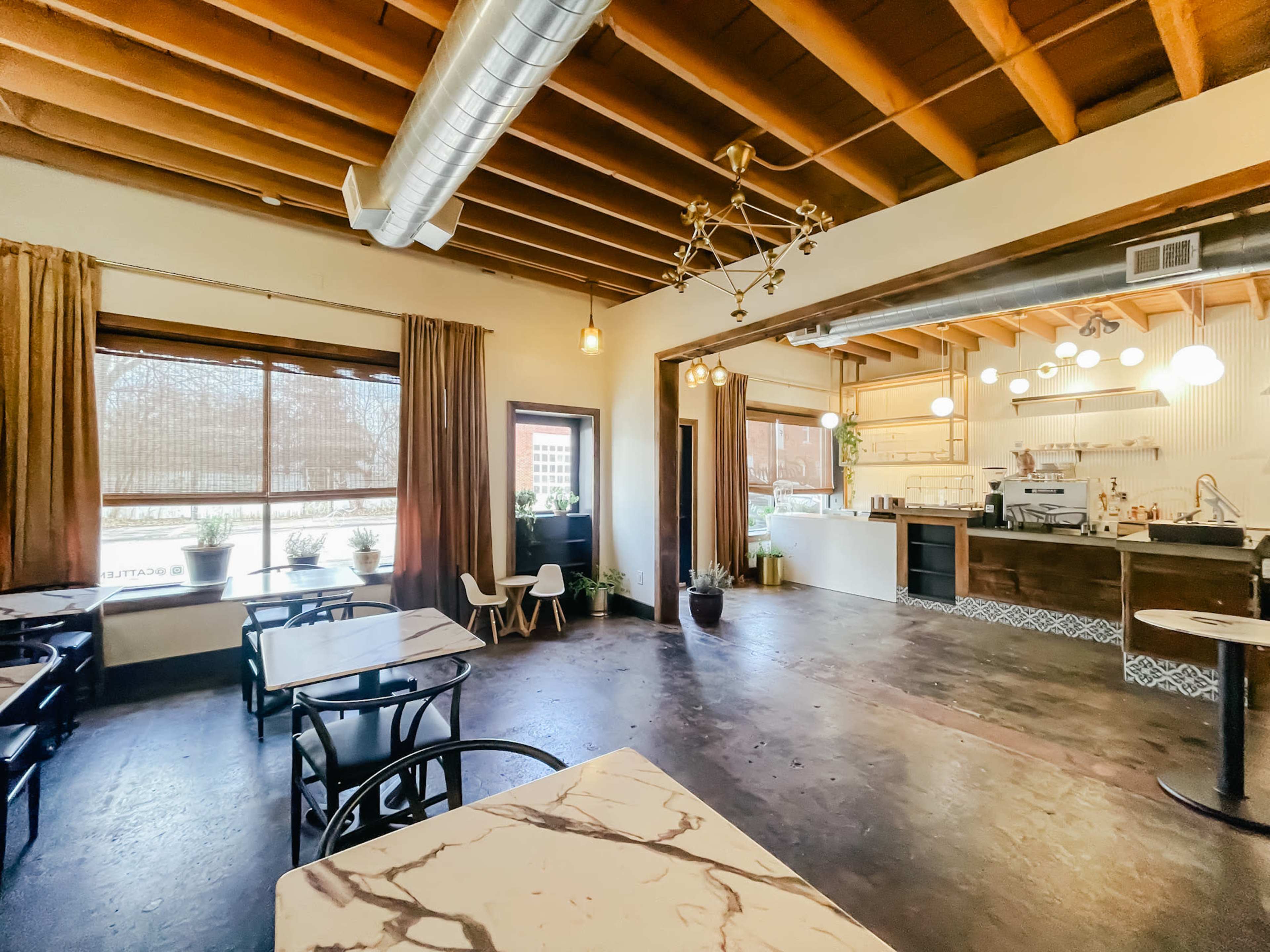 The image shows a spacious cafe with wooden beams, marble-topped tables, and large windows allowing natural light to fill the room.