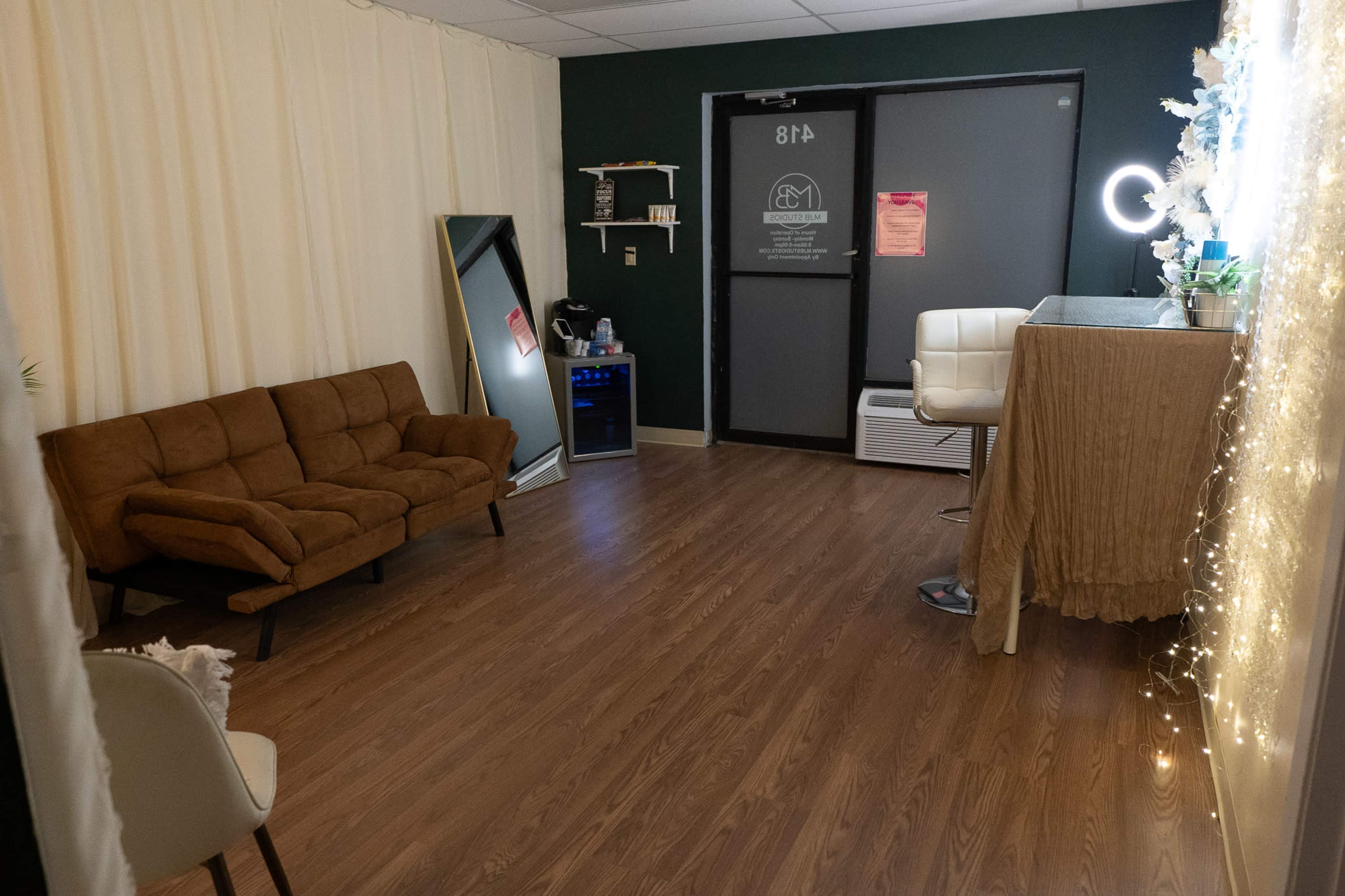 A small, softly lit room featuring a brown sofa, a mirror, a mini fridge, and a desk with decorative elements, along with a large door.