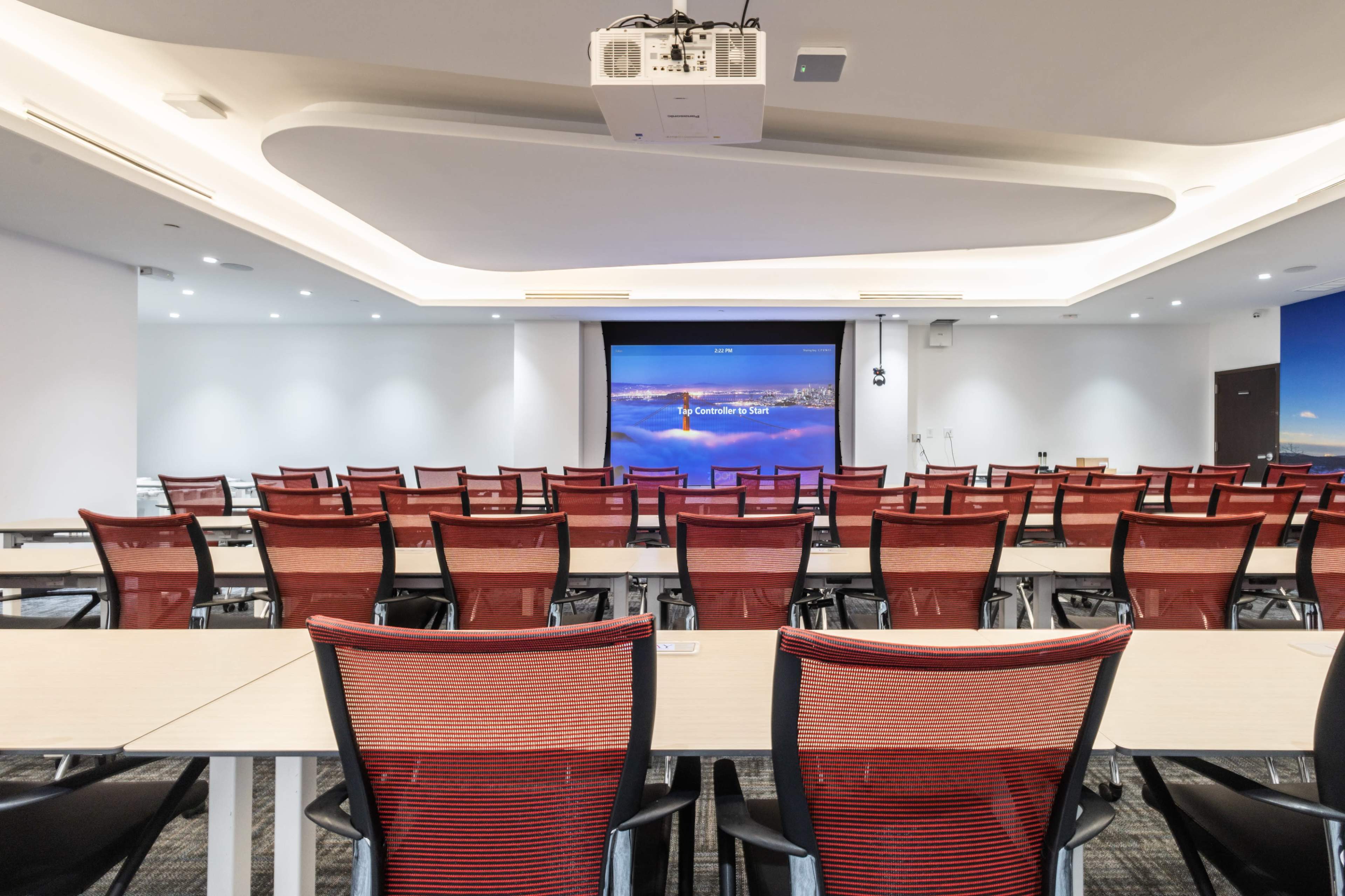 A conference room arranged with rows of red mesh chairs facing a projector screen at the front.