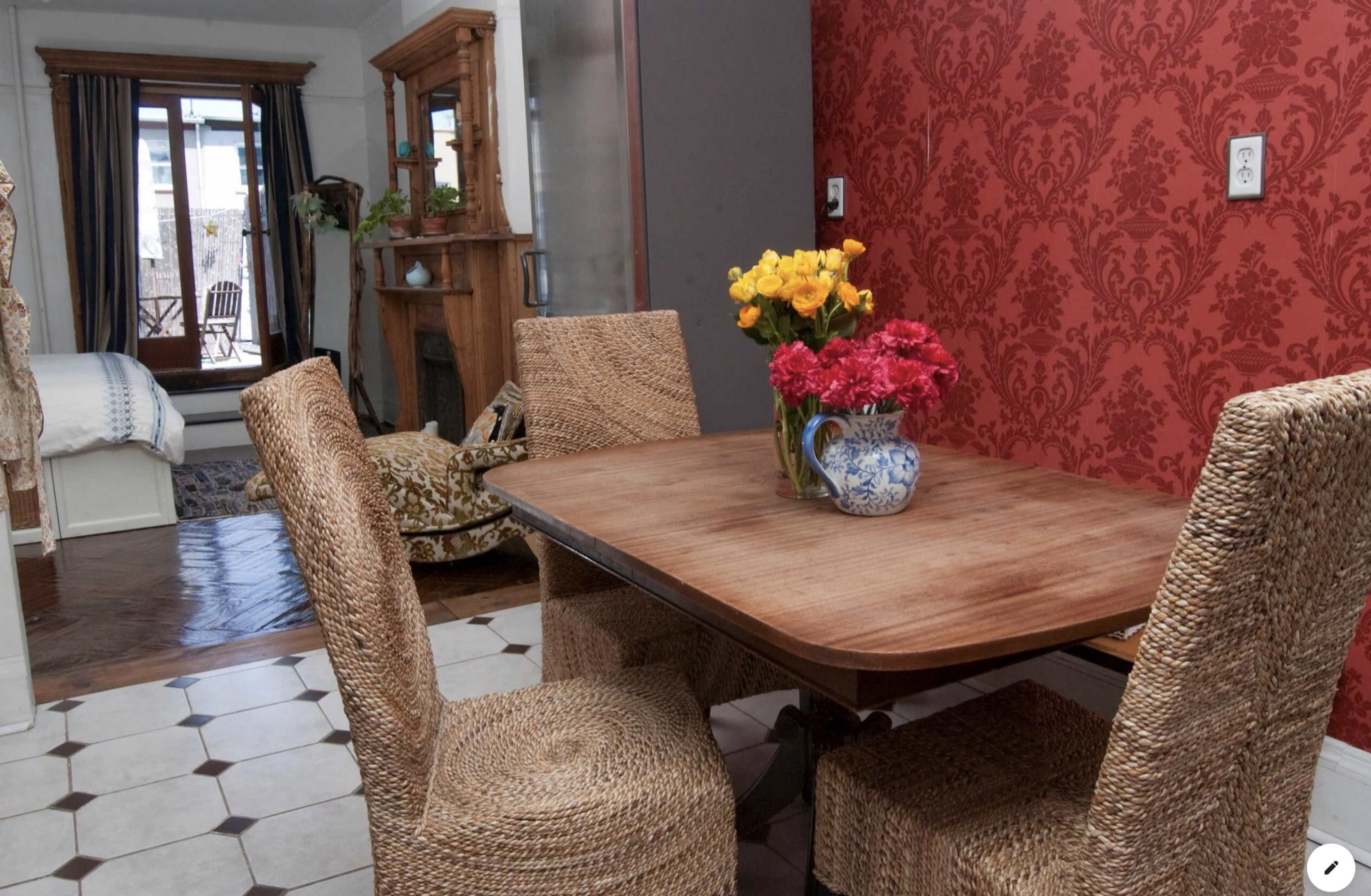 A wooden dining table with two woven chairs is set near a wall decorated with a red floral pattern, while a vase of flowers sits at the center of the table.
