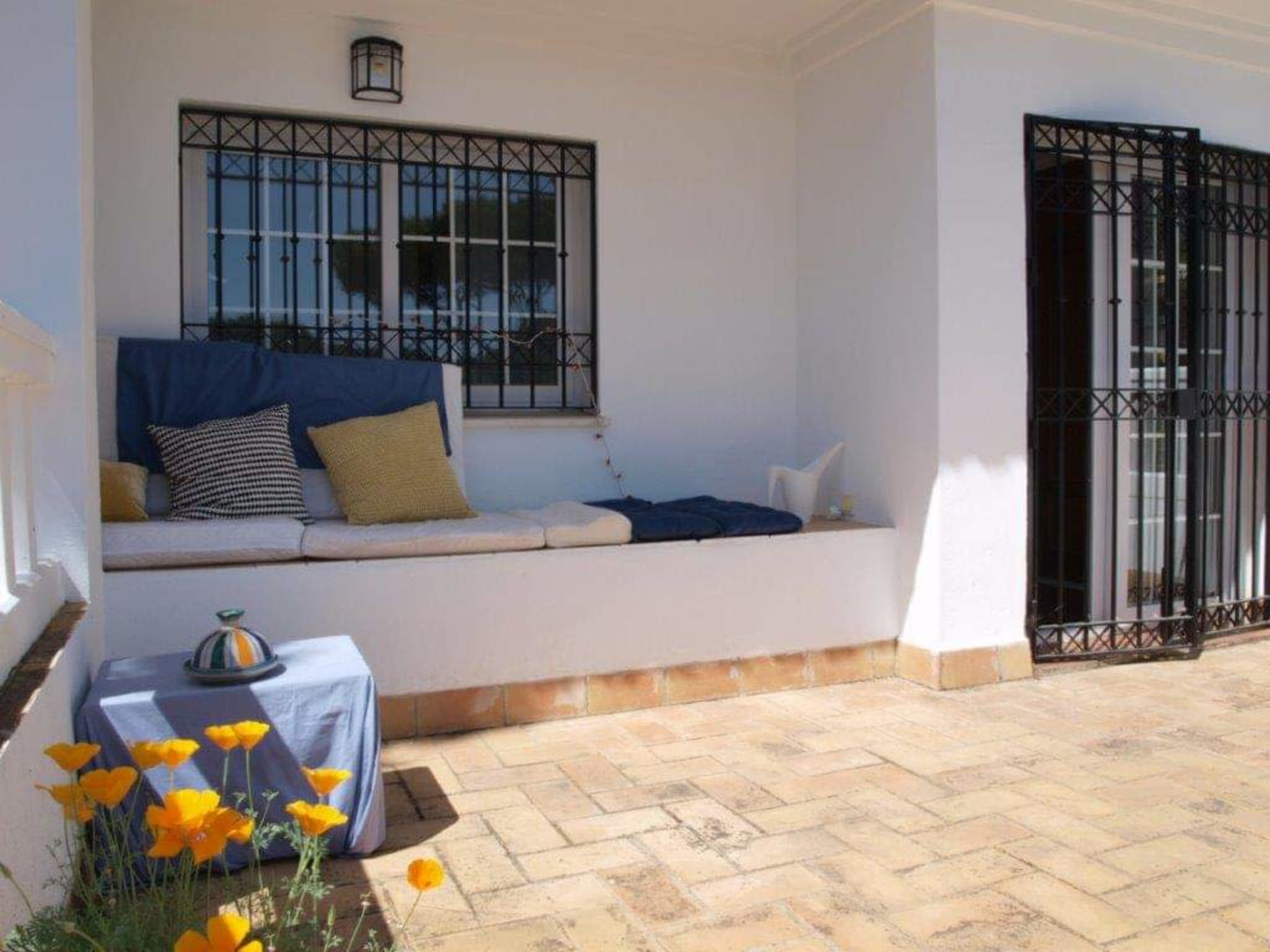 A patio features a built-in seating area with cushions, a small table, and yellow flowers in front of a white wall with black security bars on the windows and door.