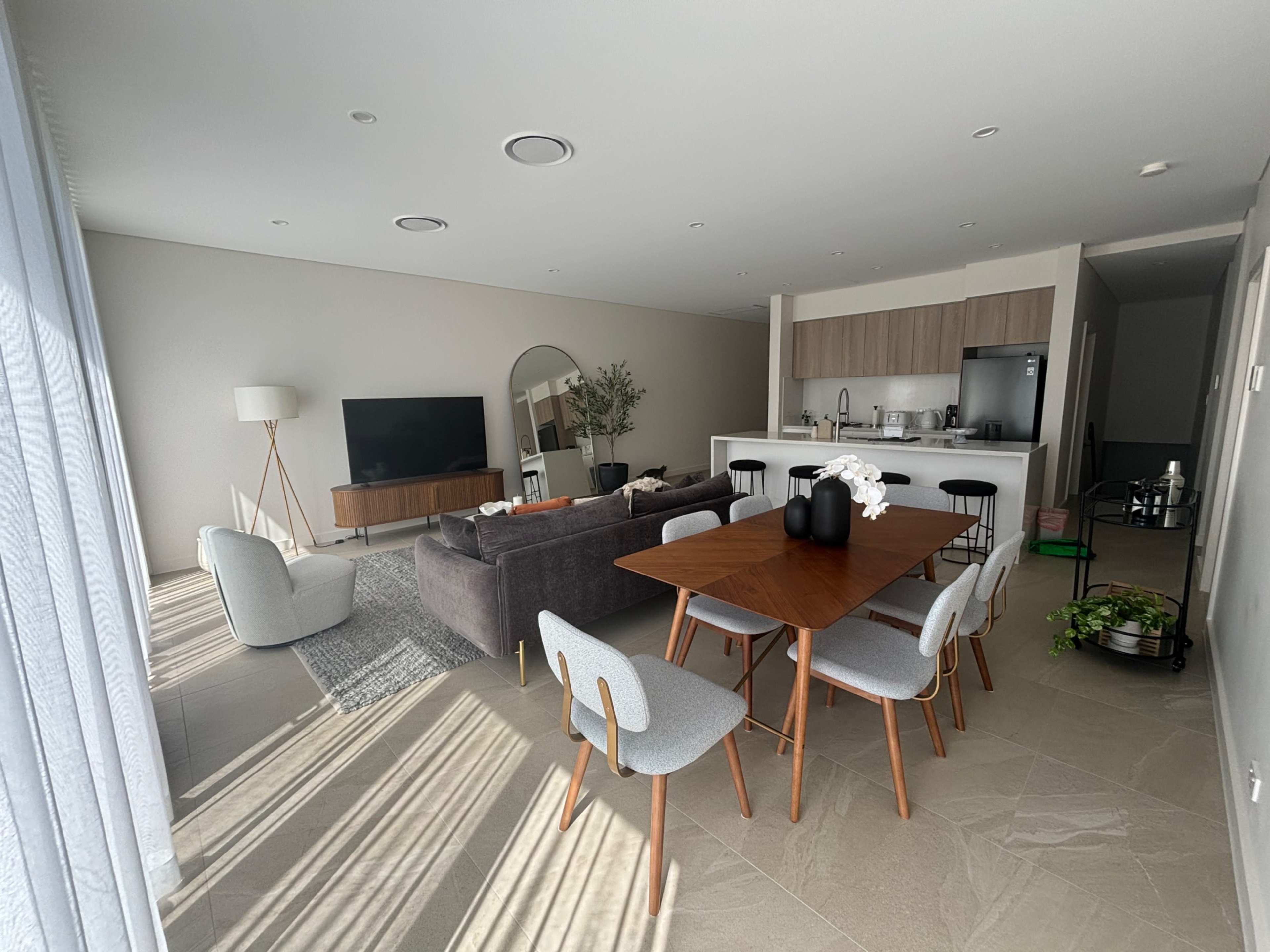 A modern open-concept living and dining area with a large dining table, a sofa, a television, and a kitchen in the background.