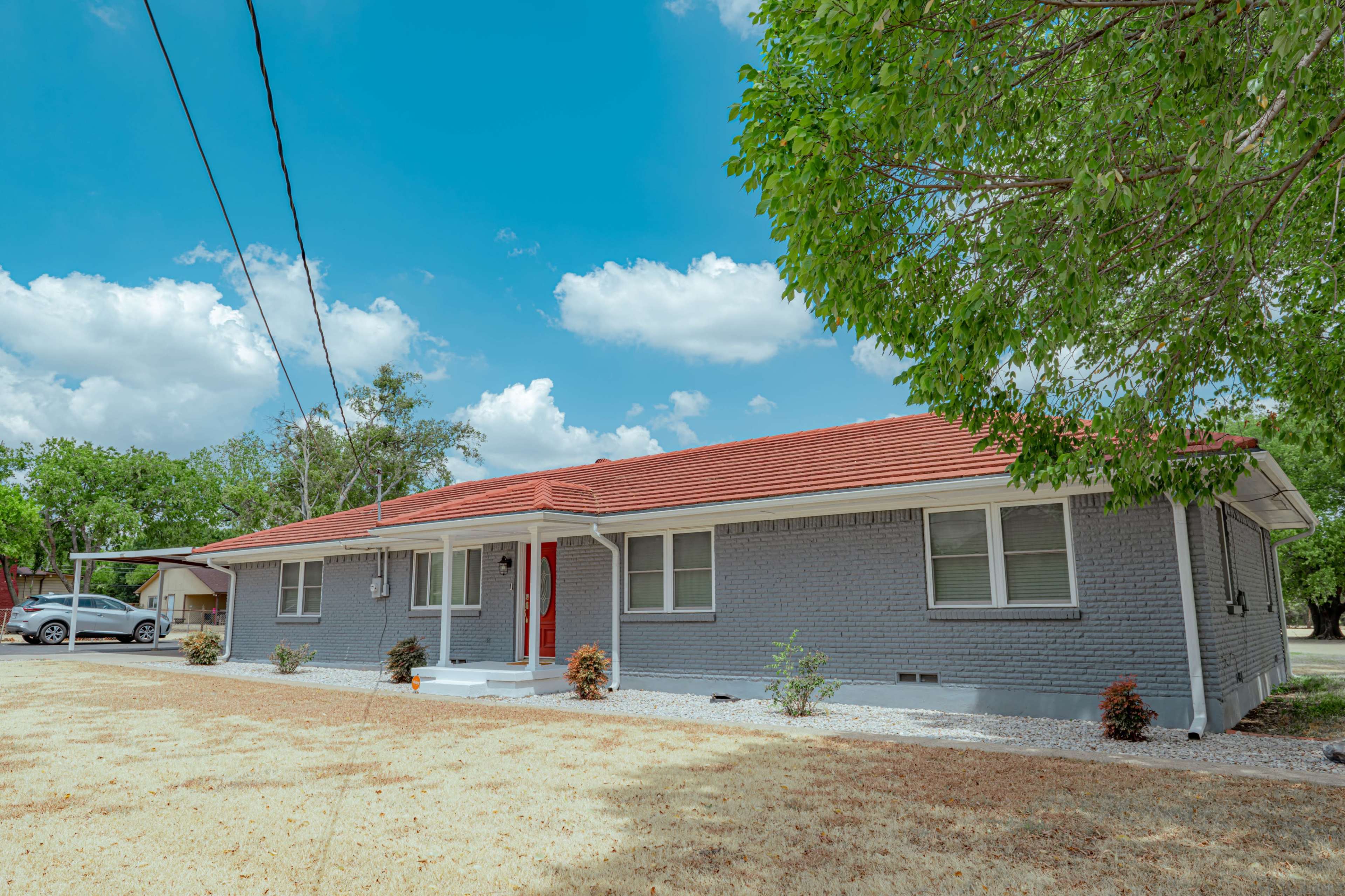 A single-story grey brick house with a red roof, surrounded by sparse vegetation and a clear sky.