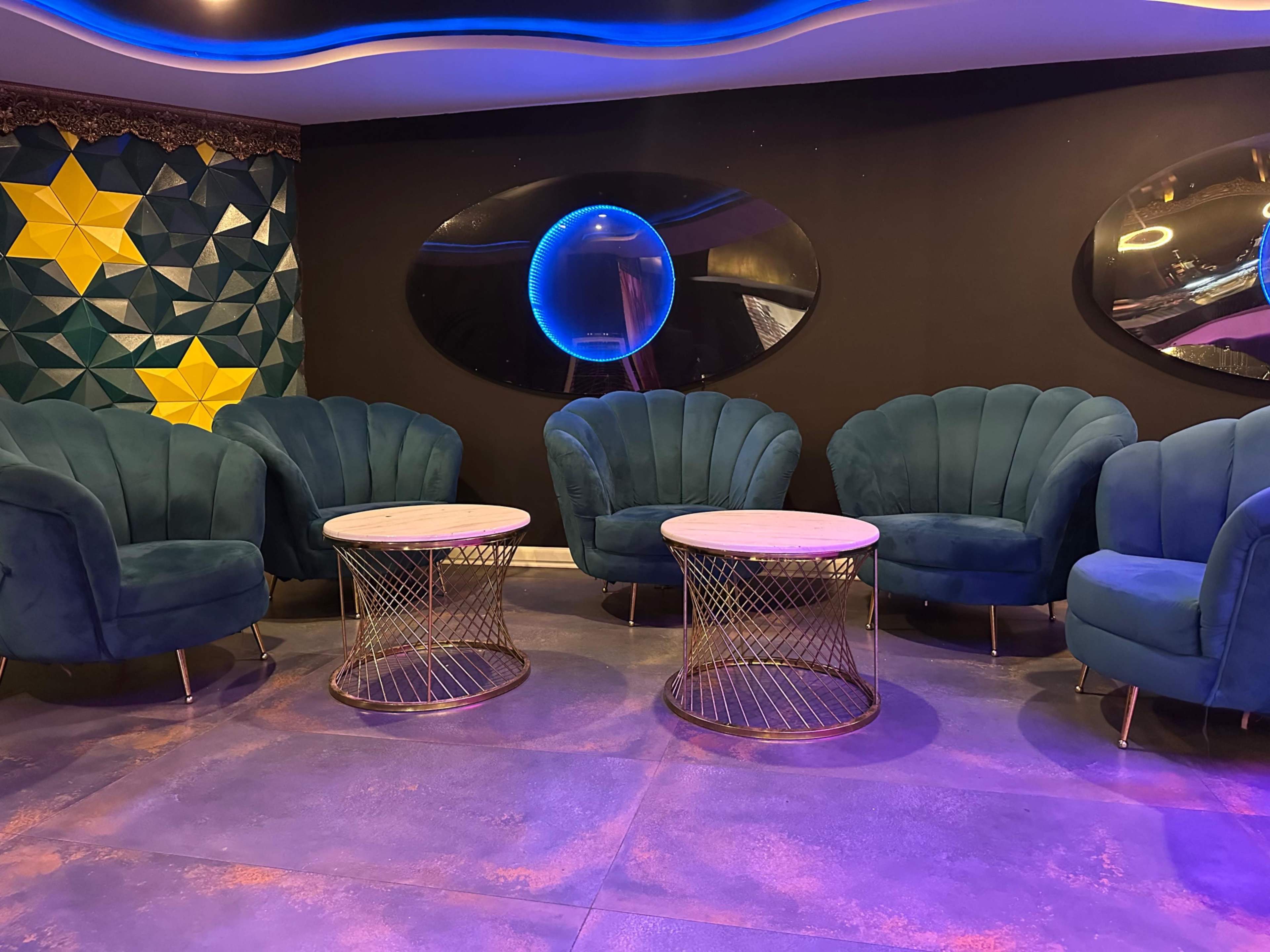 The image shows a modern lounge area featuring plush teal chairs arranged around two circular glass-top tables, against a backdrop of geometric wall patterns.