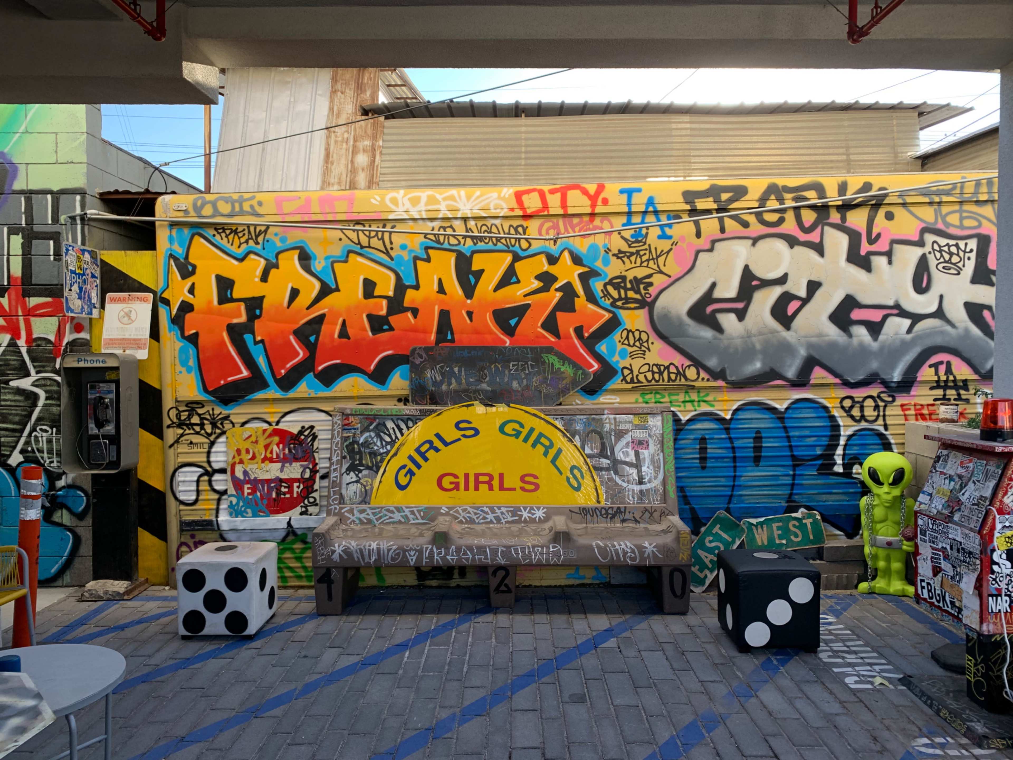 A vibrant outdoor area with a graffiti-covered wall, a yellow bench labeled "GIRLS GIRLS GIRLS," and decorations including a green alien and oversized dice.
