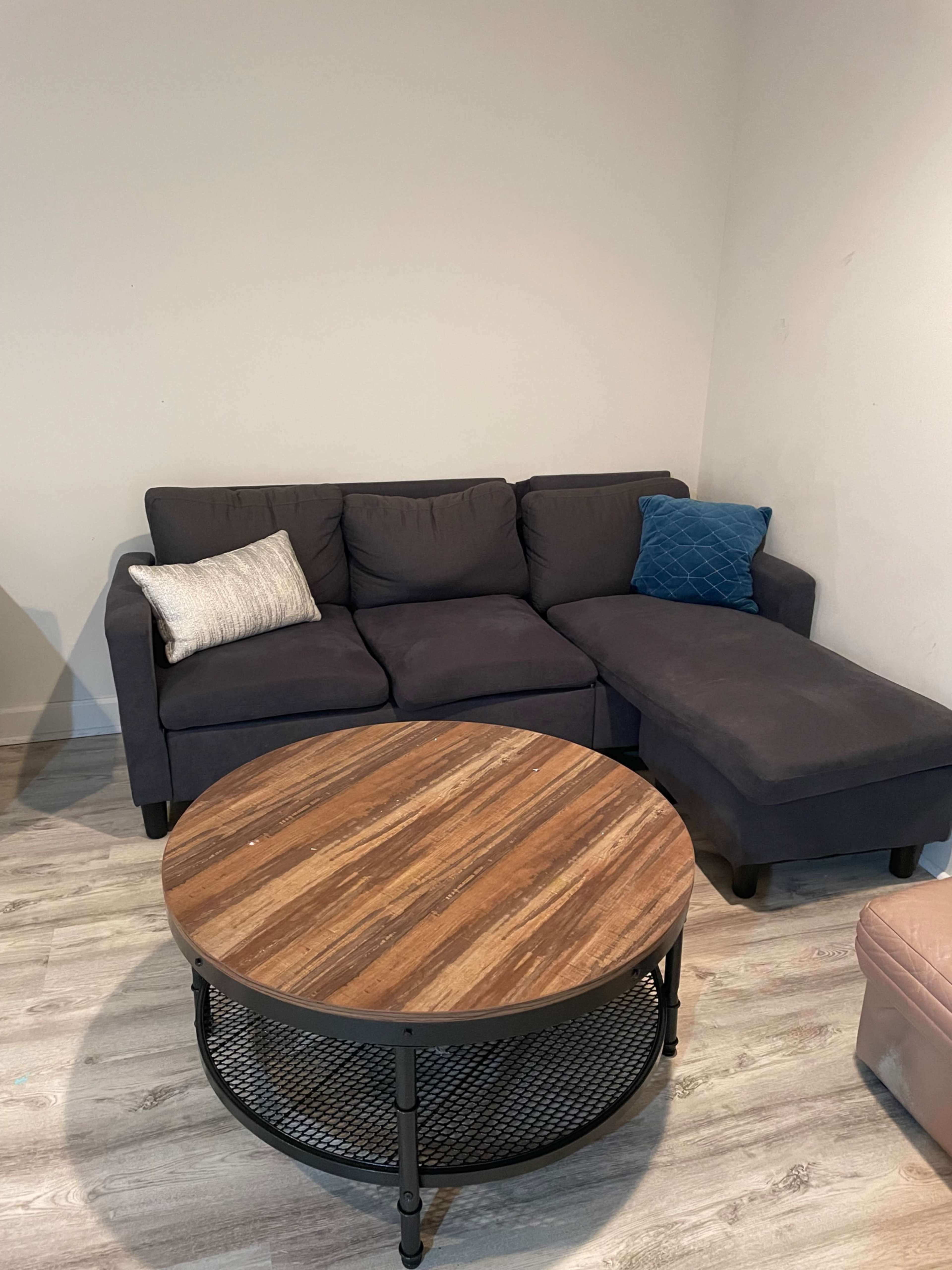The image shows a dark gray sectional sofa placed against a wall, accompanied by a round wooden coffee table with a metal base.