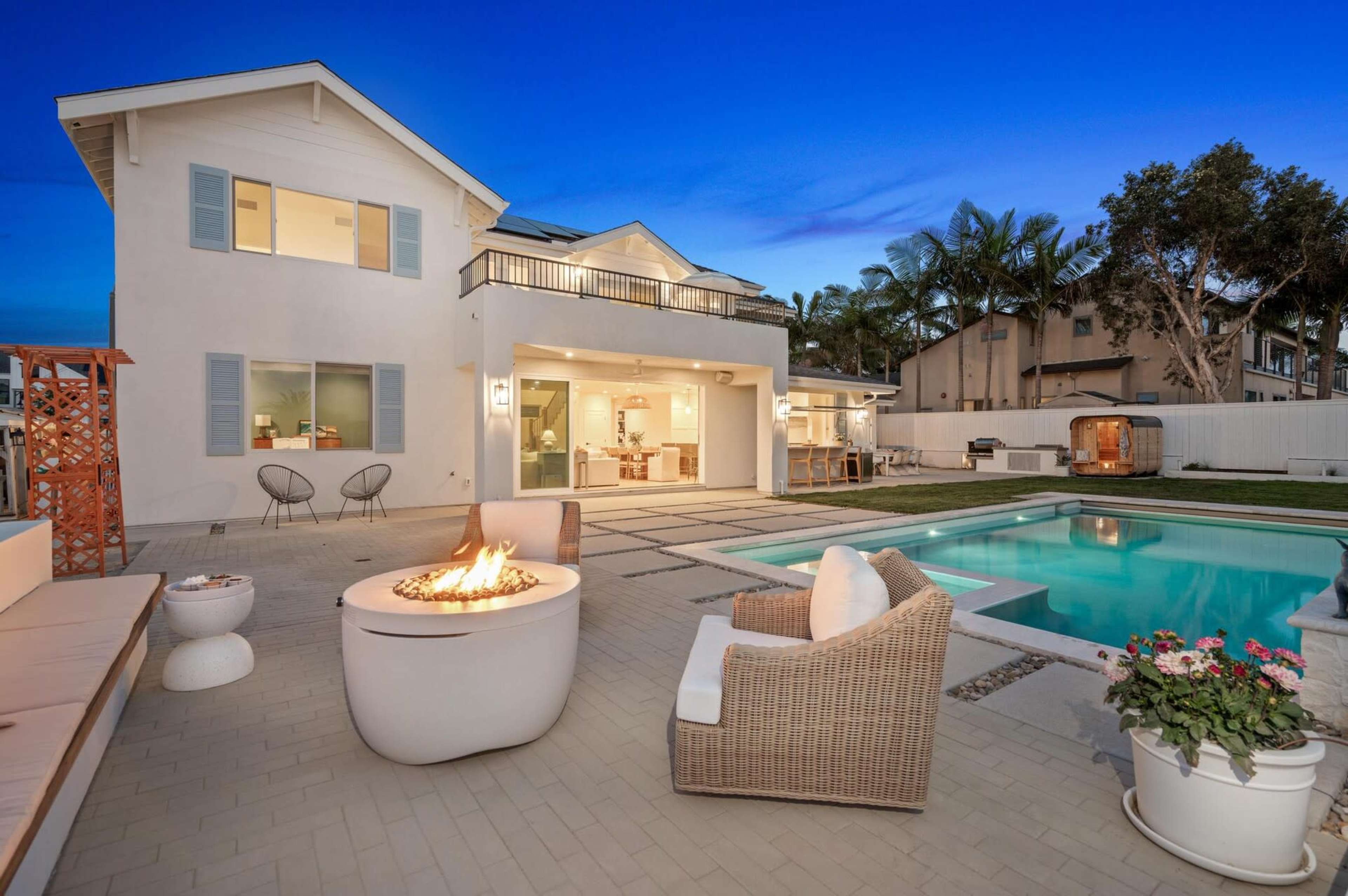 The image shows a modern two-story house with a swimming pool and a fire pit in the backyard, surrounded by patio seating and palm trees at dusk.
