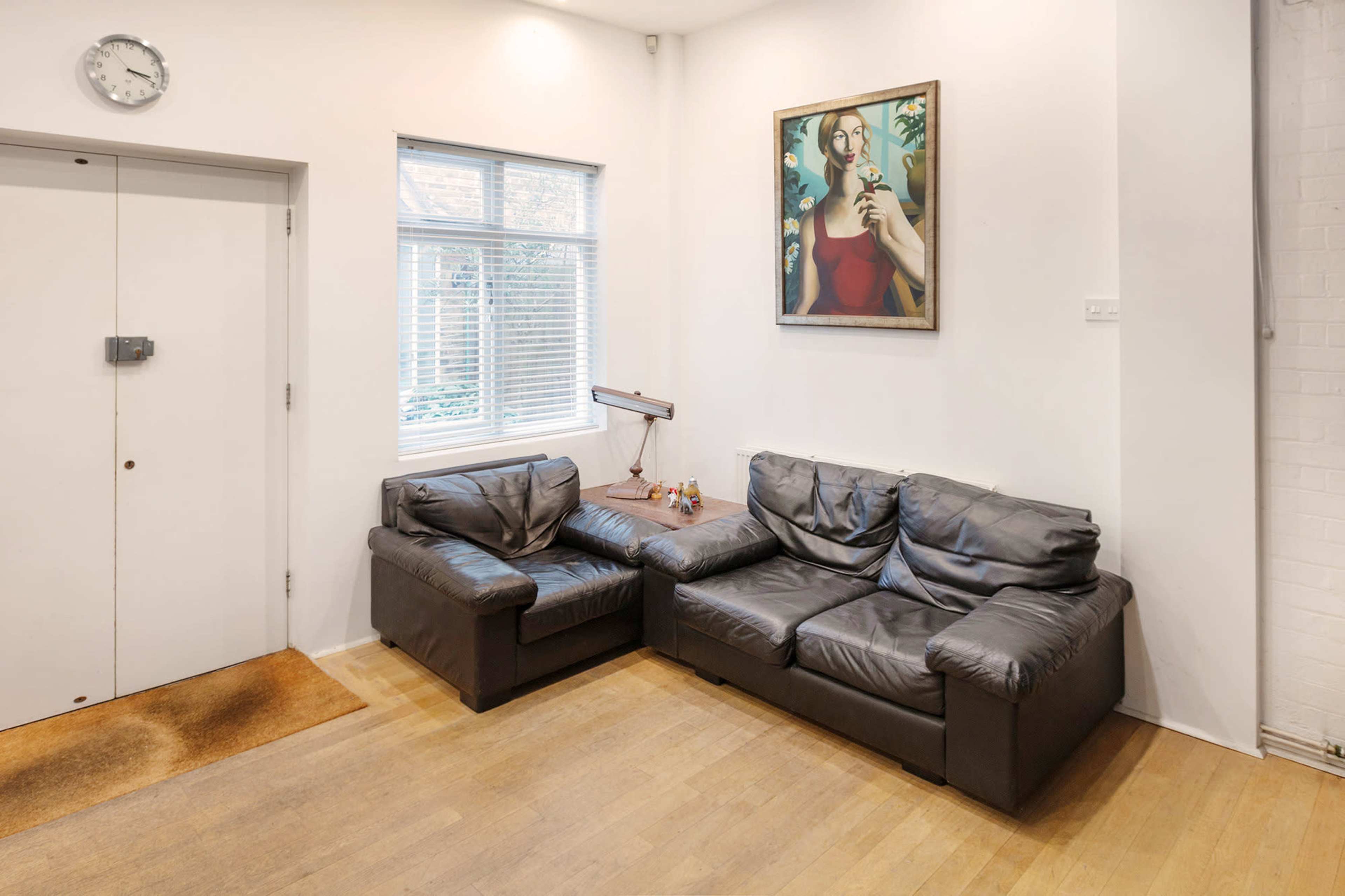 A corner of a room with a black leather sectional sofa, a wall clock, a window with blinds, and a framed painting above the couch.