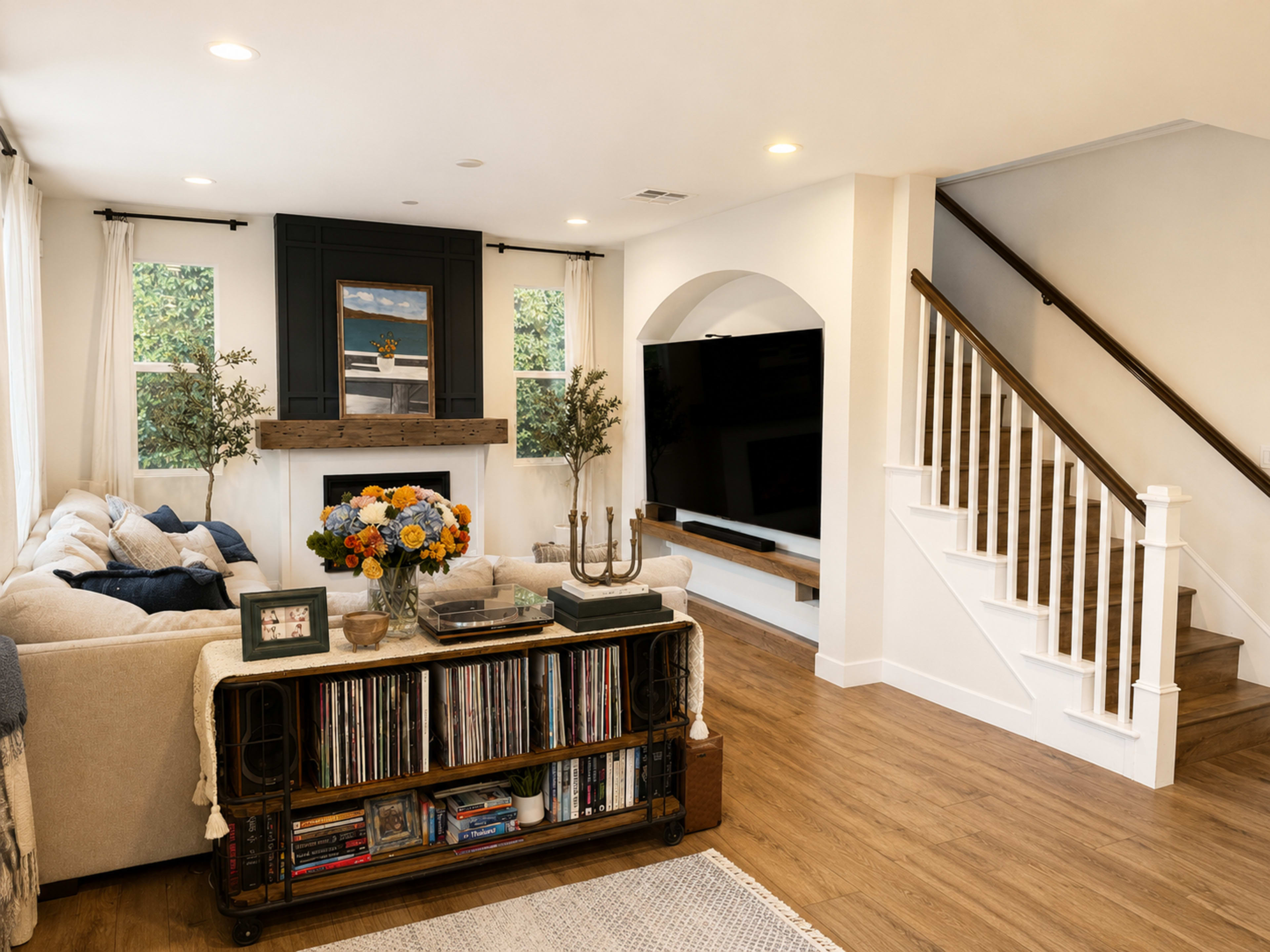 The image shows a modern living room with a sectional sofa, a wooden entertainment center, a staircase, and decorative plants.