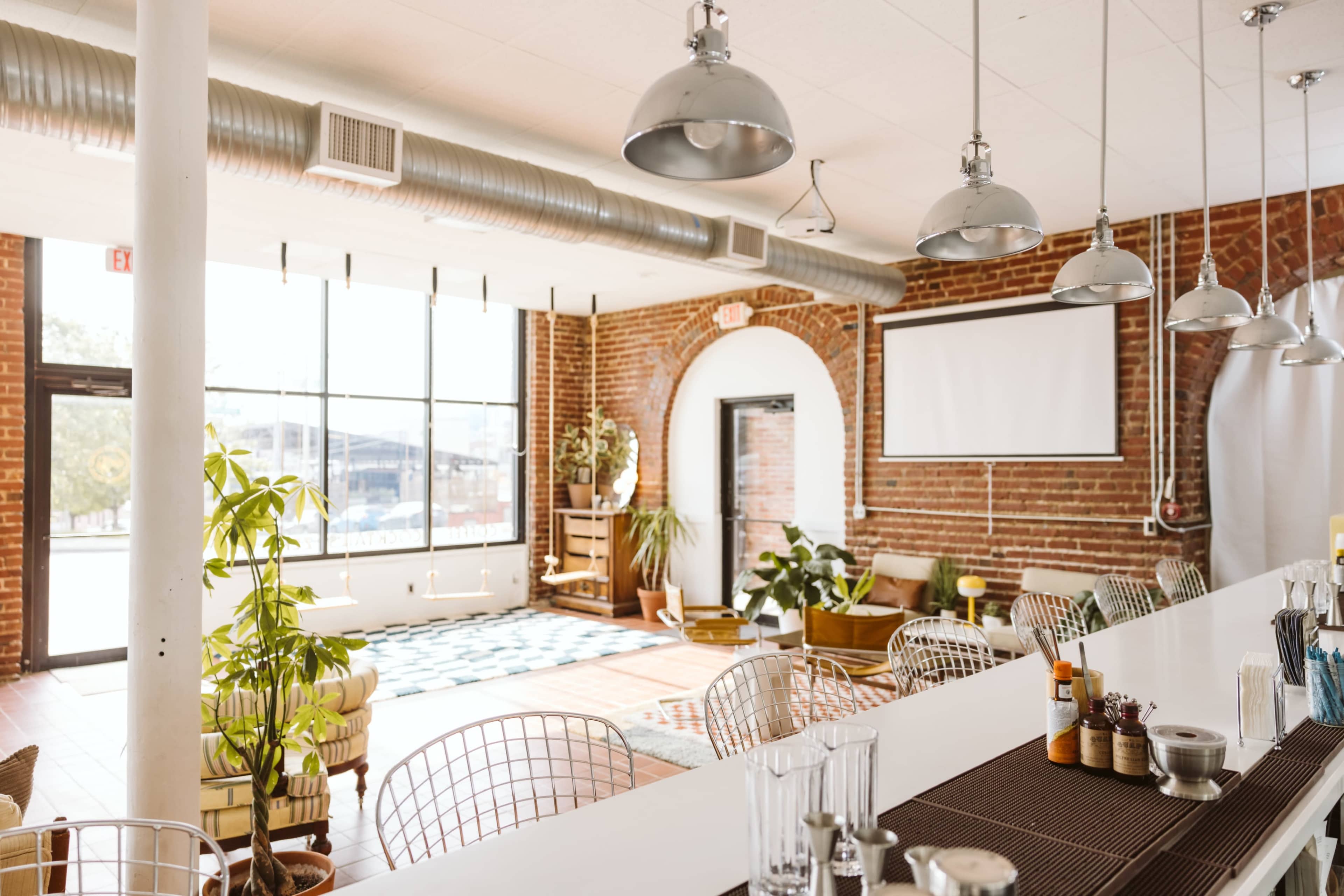 The image shows a modern interior of a cafe with large windows, exposed brick walls, and minimalist furniture, including bar stools and plants.