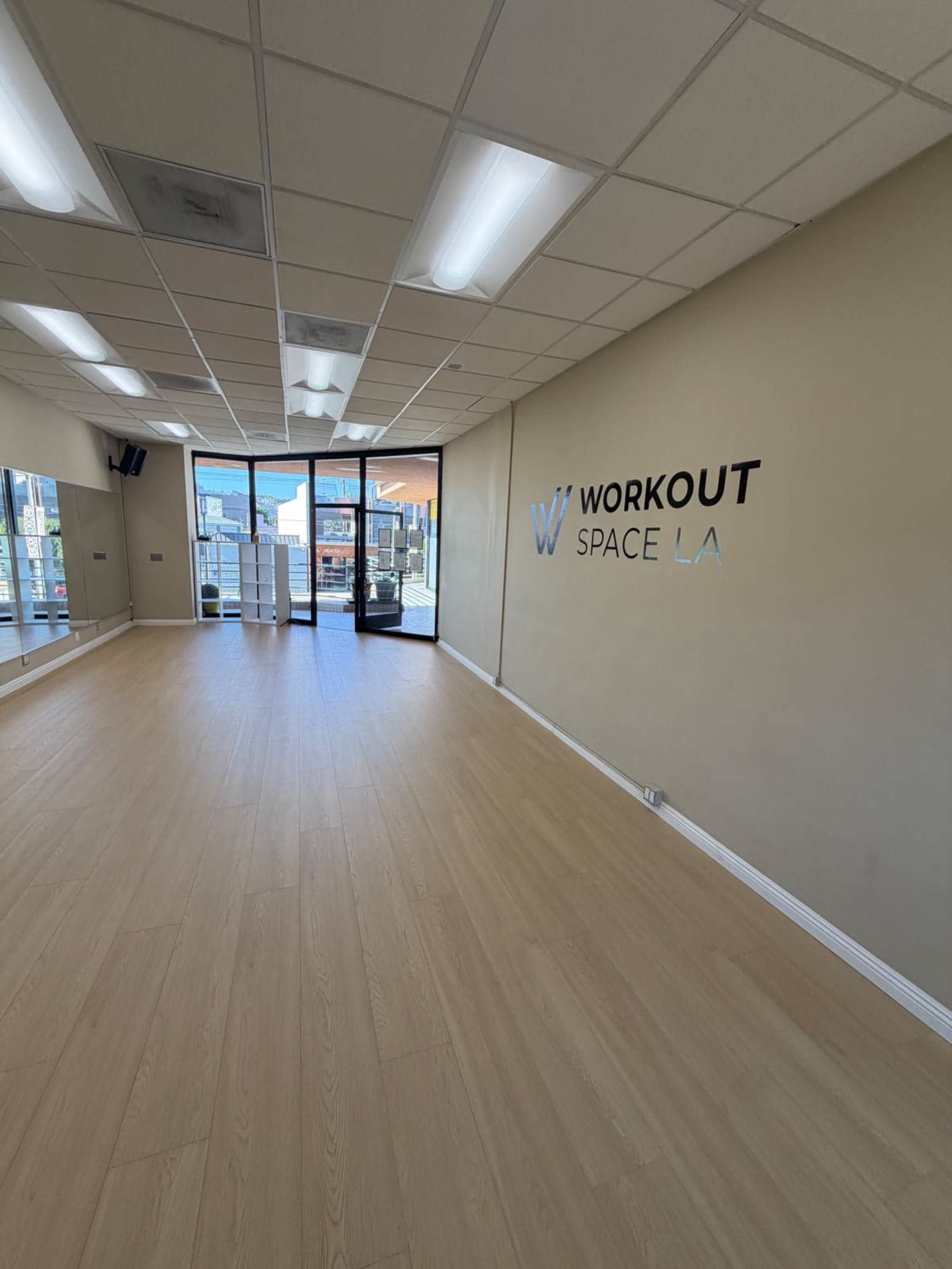The image shows a spacious, well-lit room with hardwood flooring and large windows, featuring the text "WORKOUT SPACE LA" on one wall.