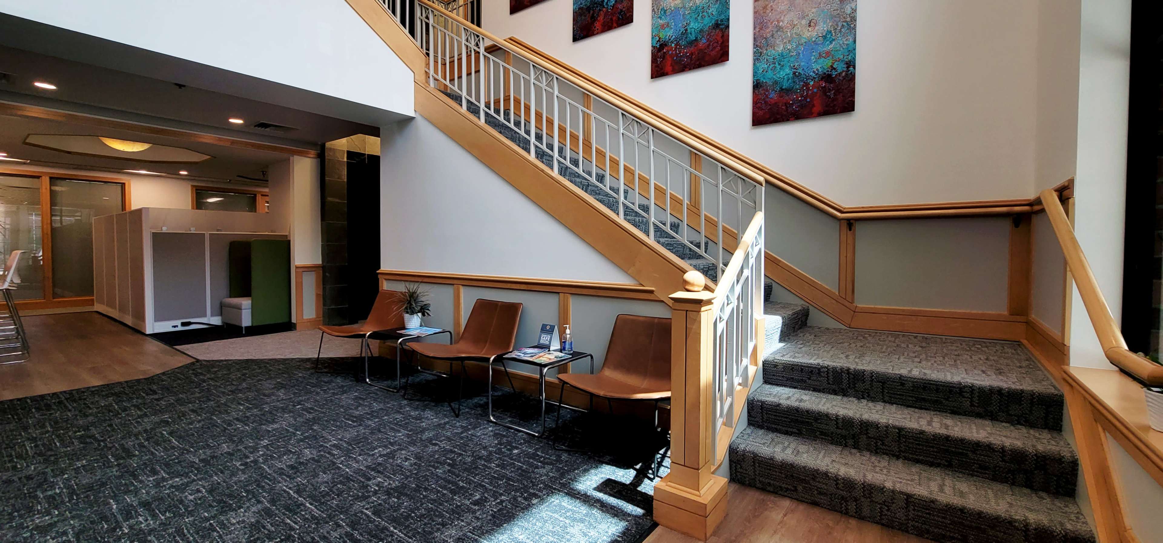 A well-lit interior space featuring a wooden staircase with railing, a seating area with modern chairs, and colorful artwork on the walls.