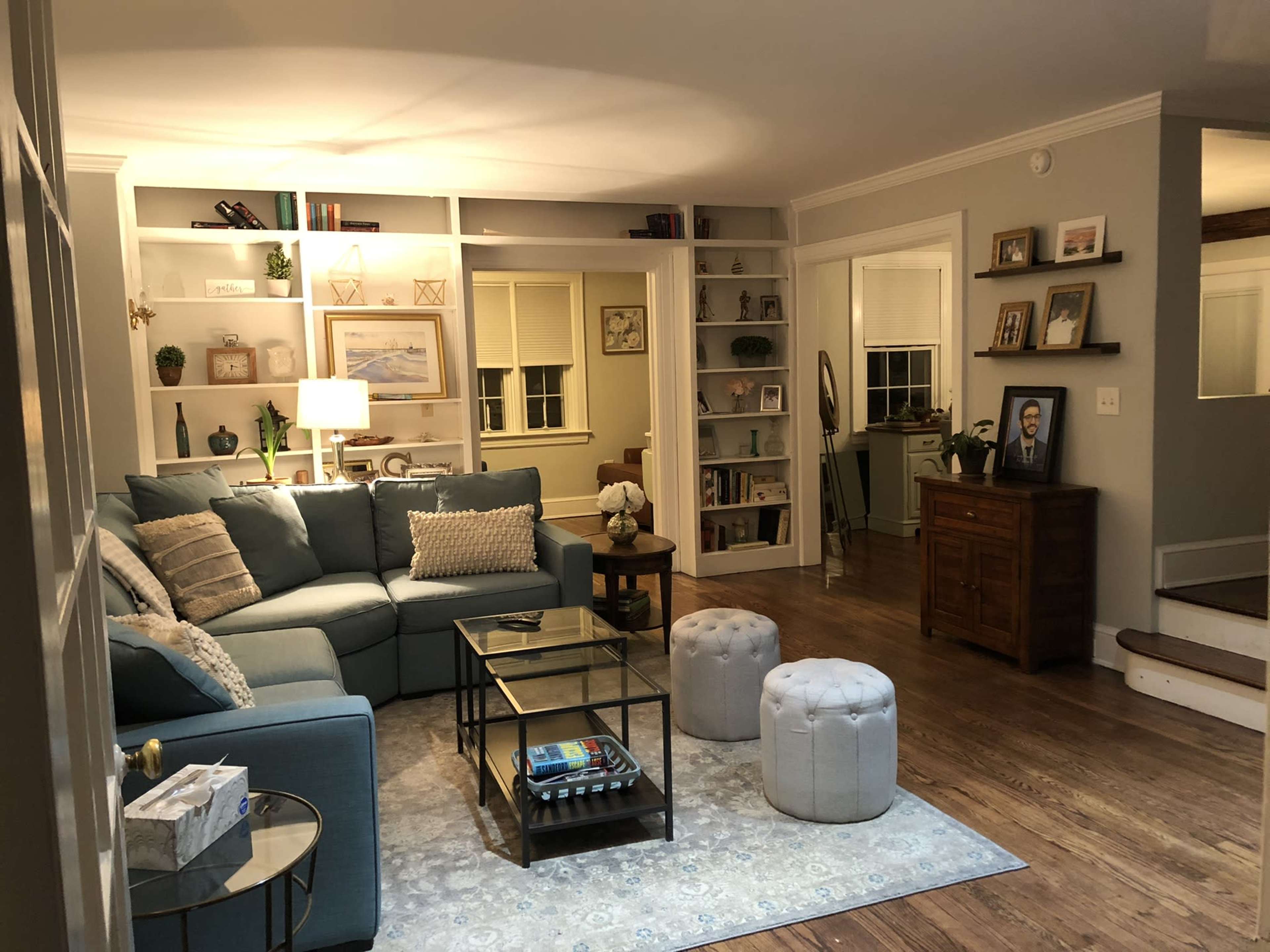 A cozy living room with a blue sectional sofa, a glass coffee table, and light-colored poufs, surrounded by bookshelves and warm wooden flooring.