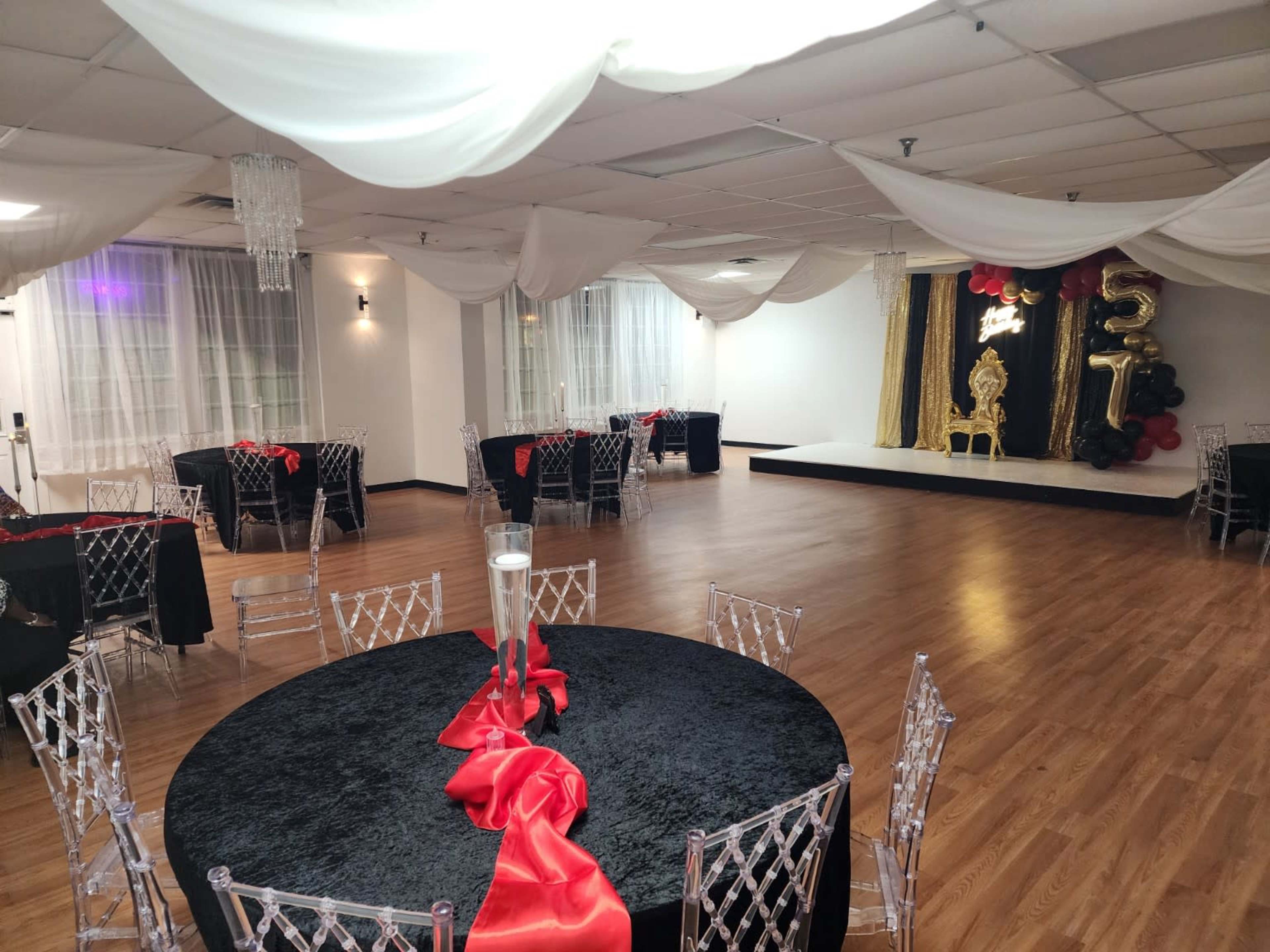 The image shows a spacious event venue with elegantly set tables adorned in black and red, arranged around a small stage decorated with balloons and draped fabric.