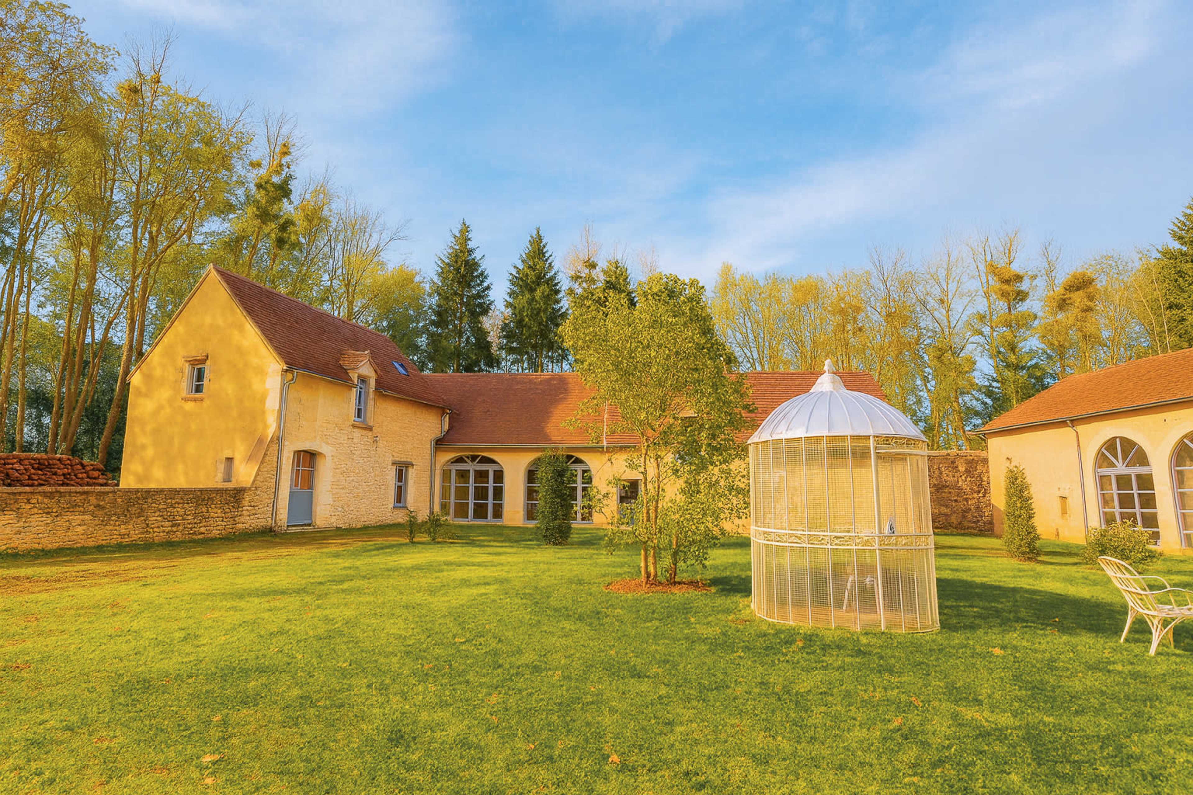 The image shows a yellow stone house with a red roof set in a green lawn, featuring a small tree and a gazebo-like structure in the garden.