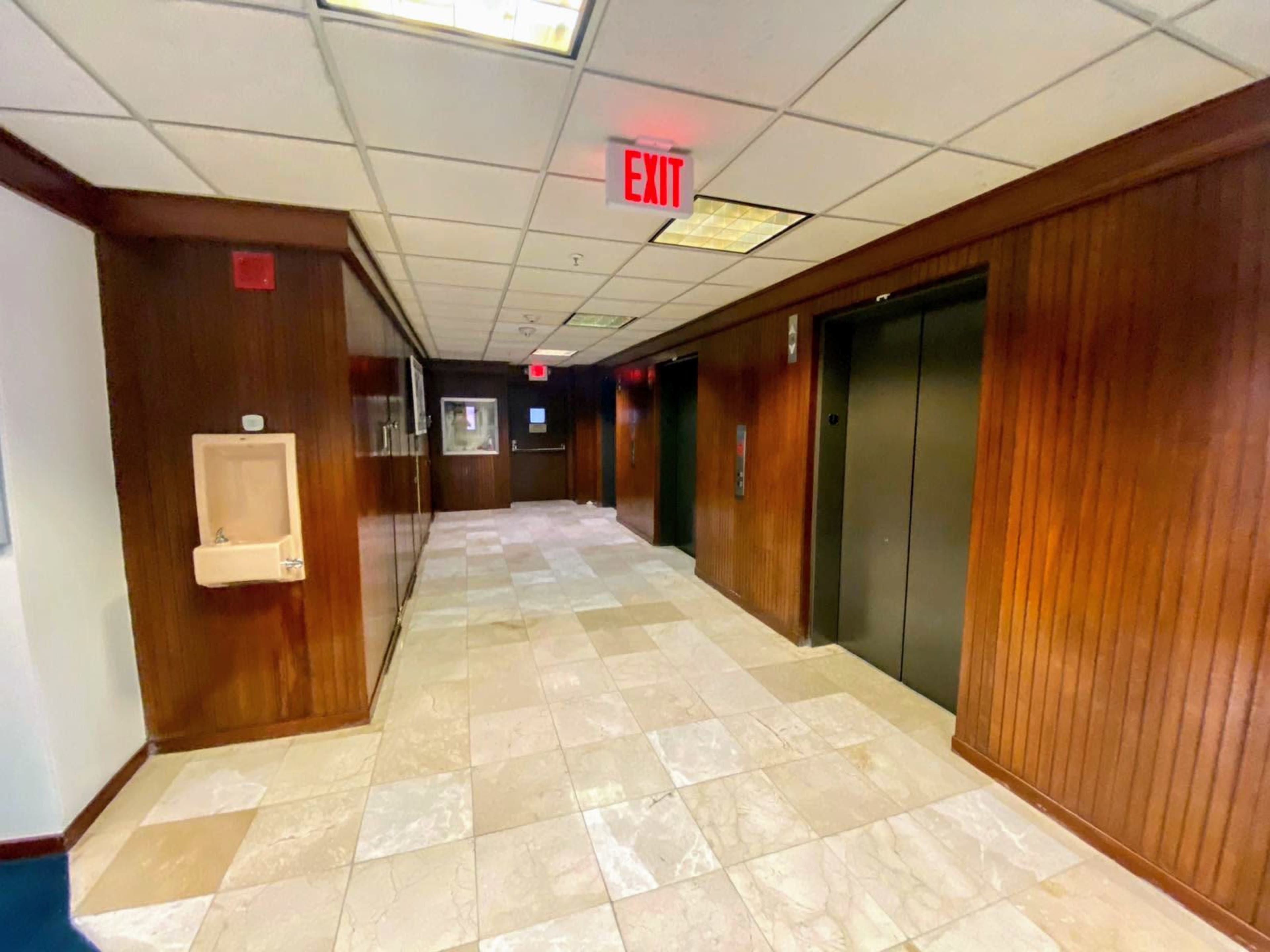 The image shows a hallway with tiled flooring, wooden paneling, and two elevators on the right side.