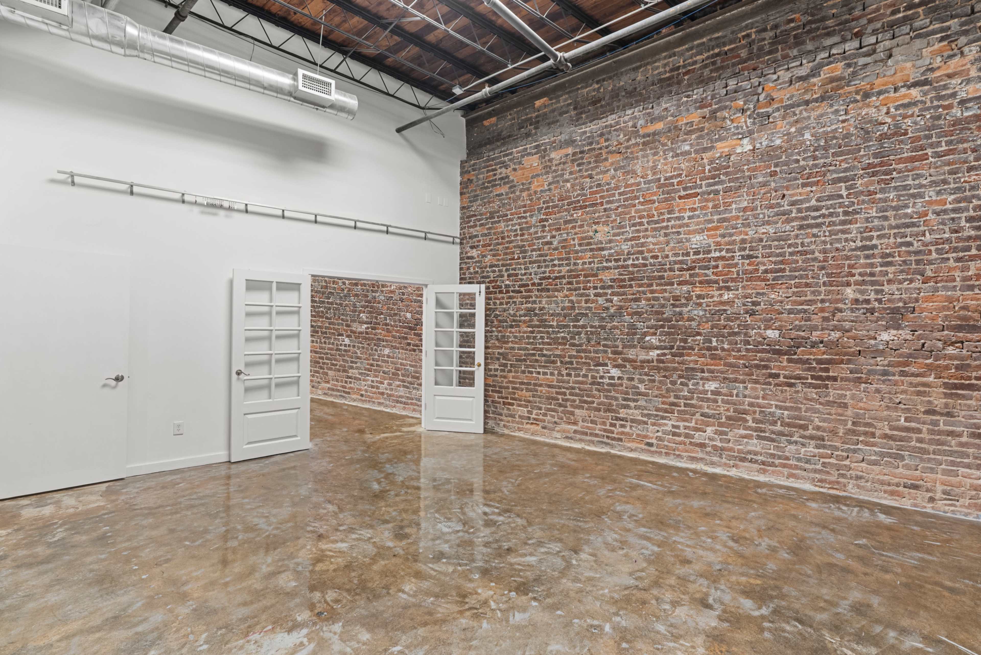 The image shows a spacious, empty room with exposed brick walls and polished concrete flooring, featuring a set of glass-paneled doors.