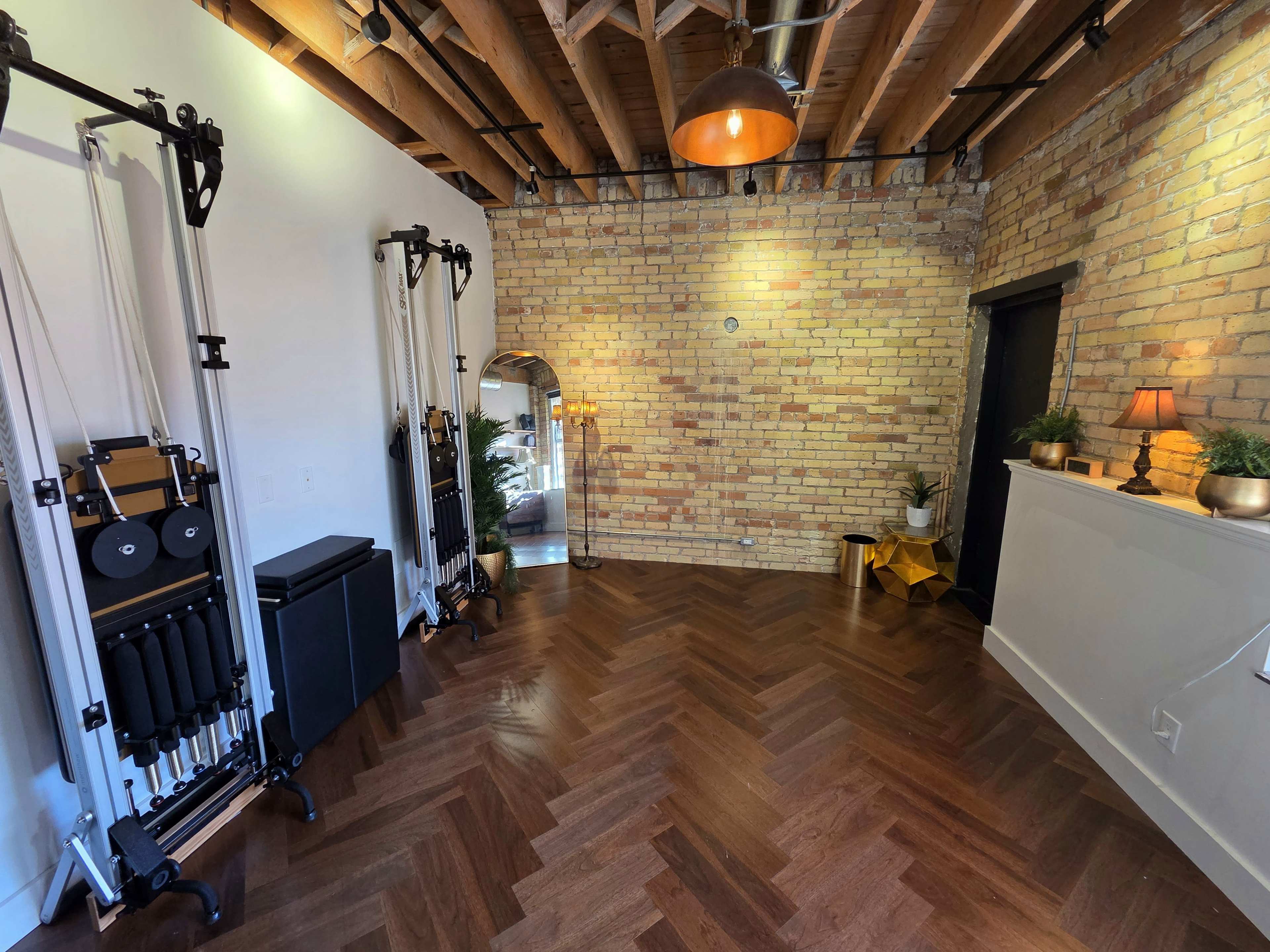 The interior of a fitness studio features wooden flooring, exposed brick walls, two reformer pilates machines on one side, and a large mirror reflecting the space.