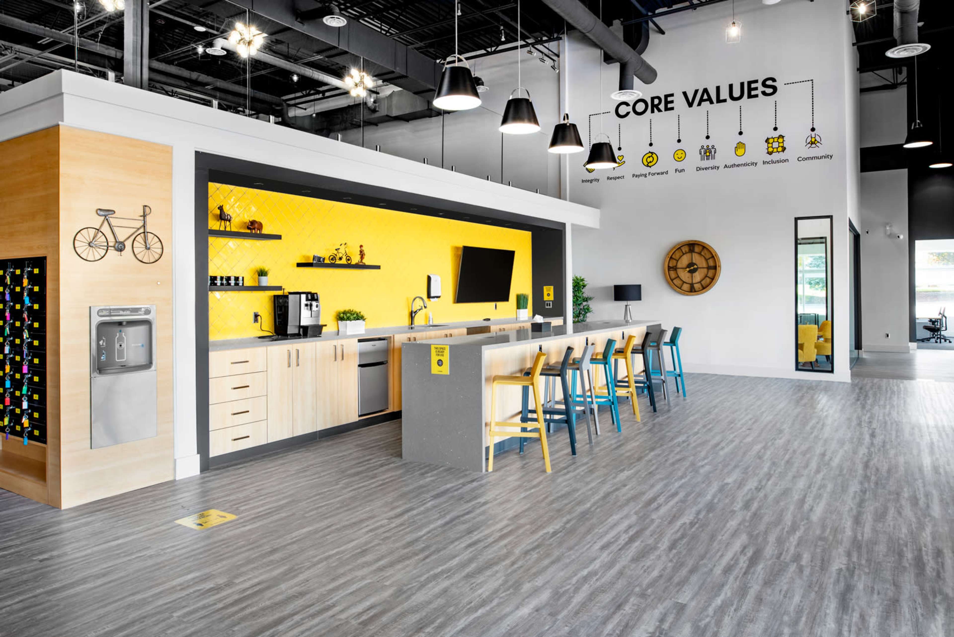 A modern kitchen area with a long bar, bright yellow accent wall, and various appliances, surrounded by colorful stools in a well-lit, open space.