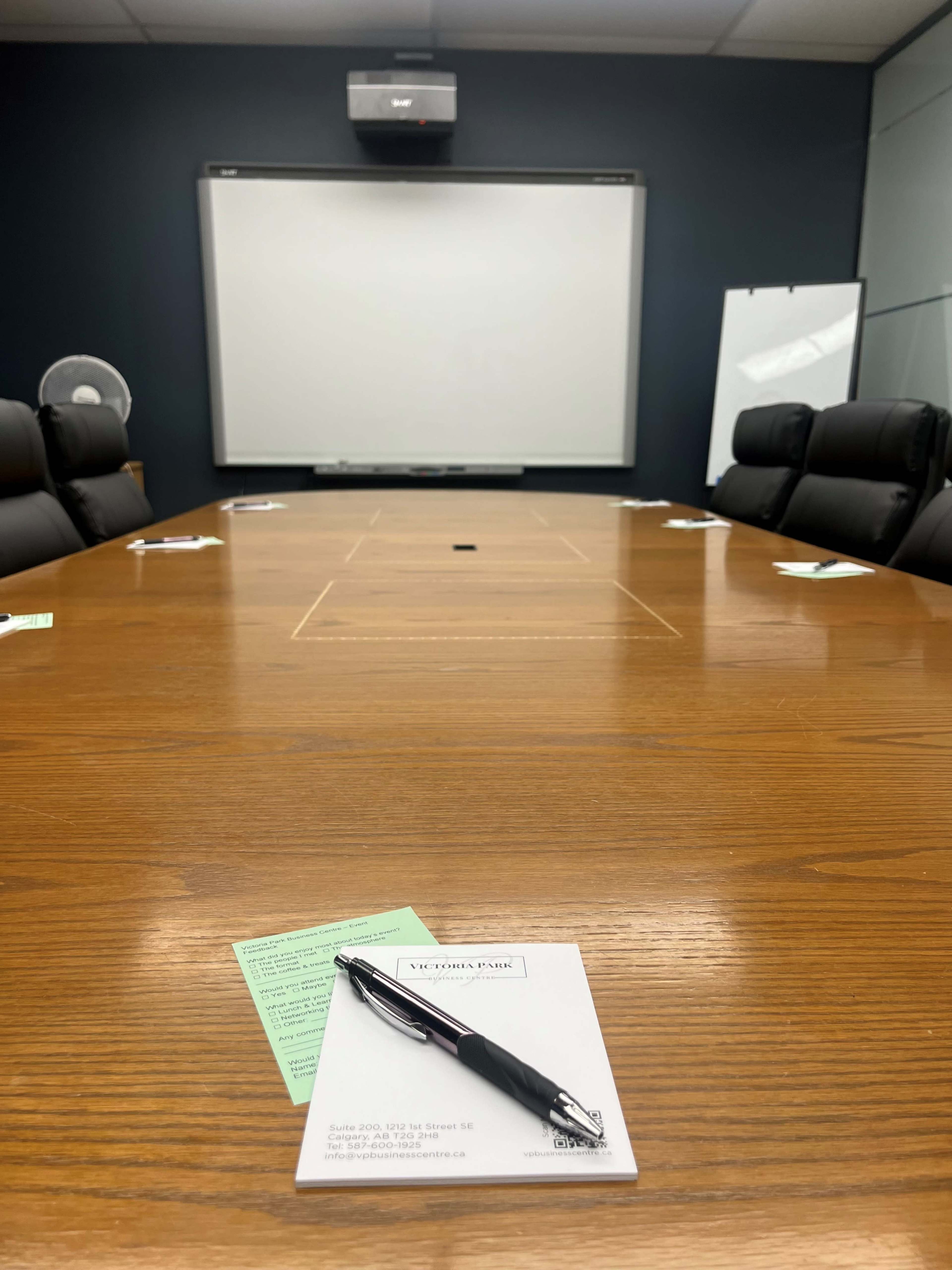 A large, wooden conference table is set up in a meeting room with a projector screen, empty chairs around it, and notepads with a pen placed at one end.