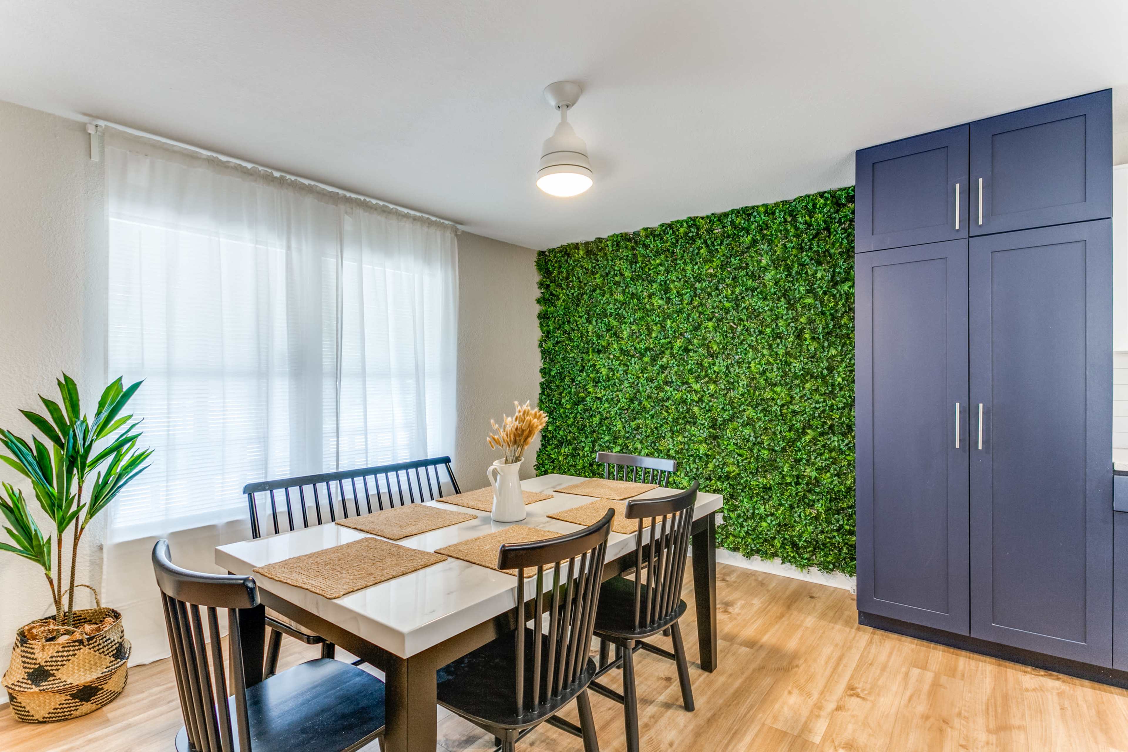 A dining area features a table with black chairs, a vase of dried flowers, and a green wall of artificial plants, alongside a dark blue cabinet and a large window with sheer curtains.