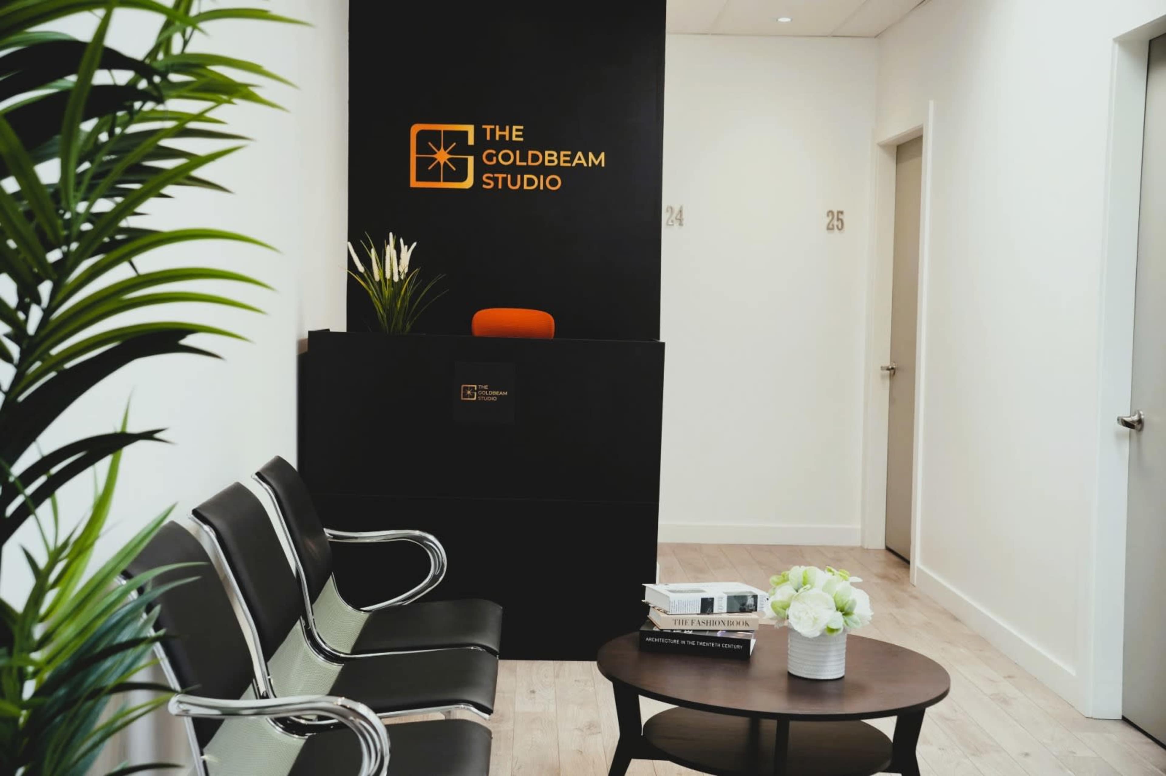 The image shows a modern reception area for "The Goldbeam Studio," featuring a black front desk, a waiting area with silver chairs, and a round table with a floral arrangement.