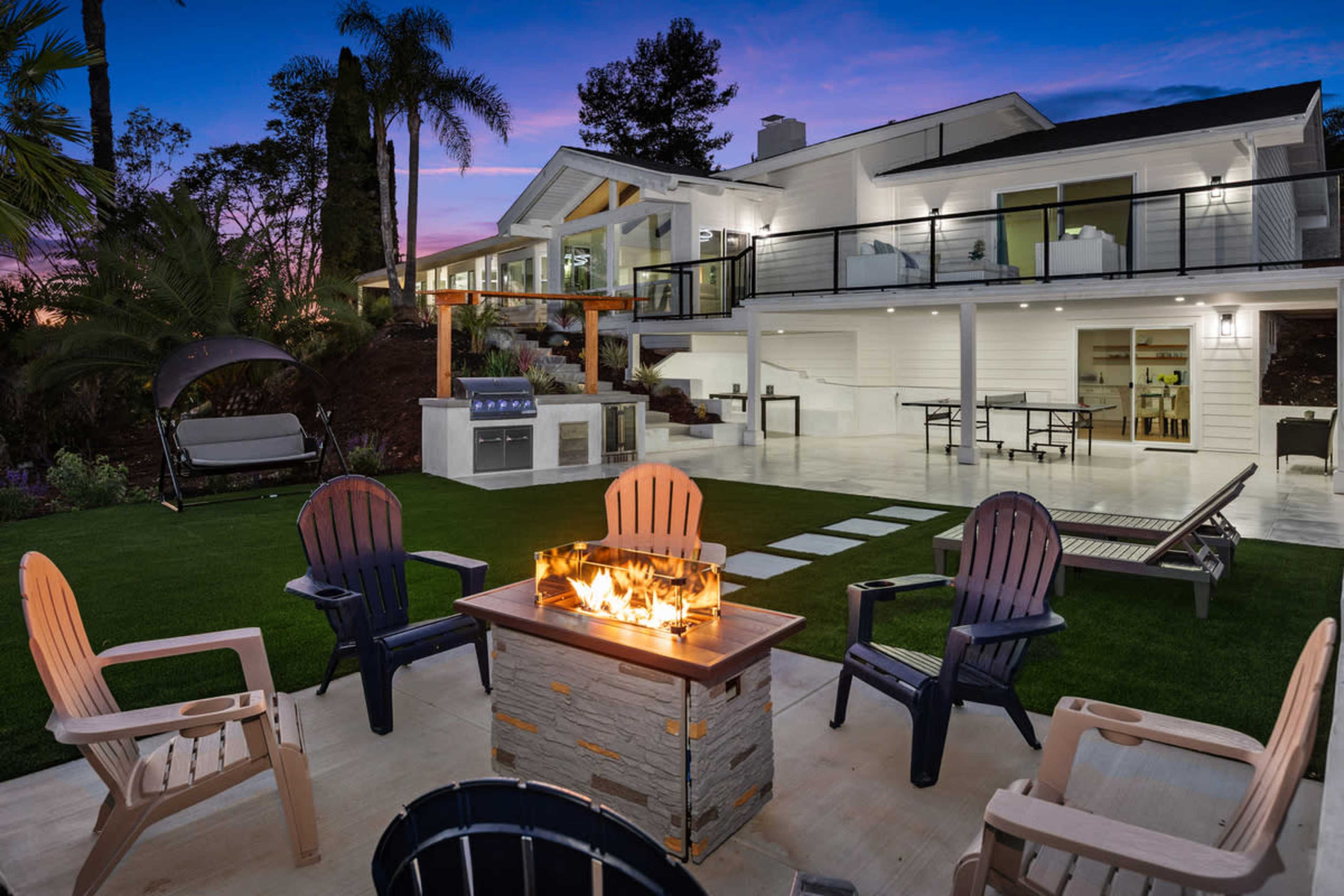 The image shows a modern outdoor living space with a fire pit surrounded by chairs, a barbecue area, and a two-story house in the background, set against a twilight sky.