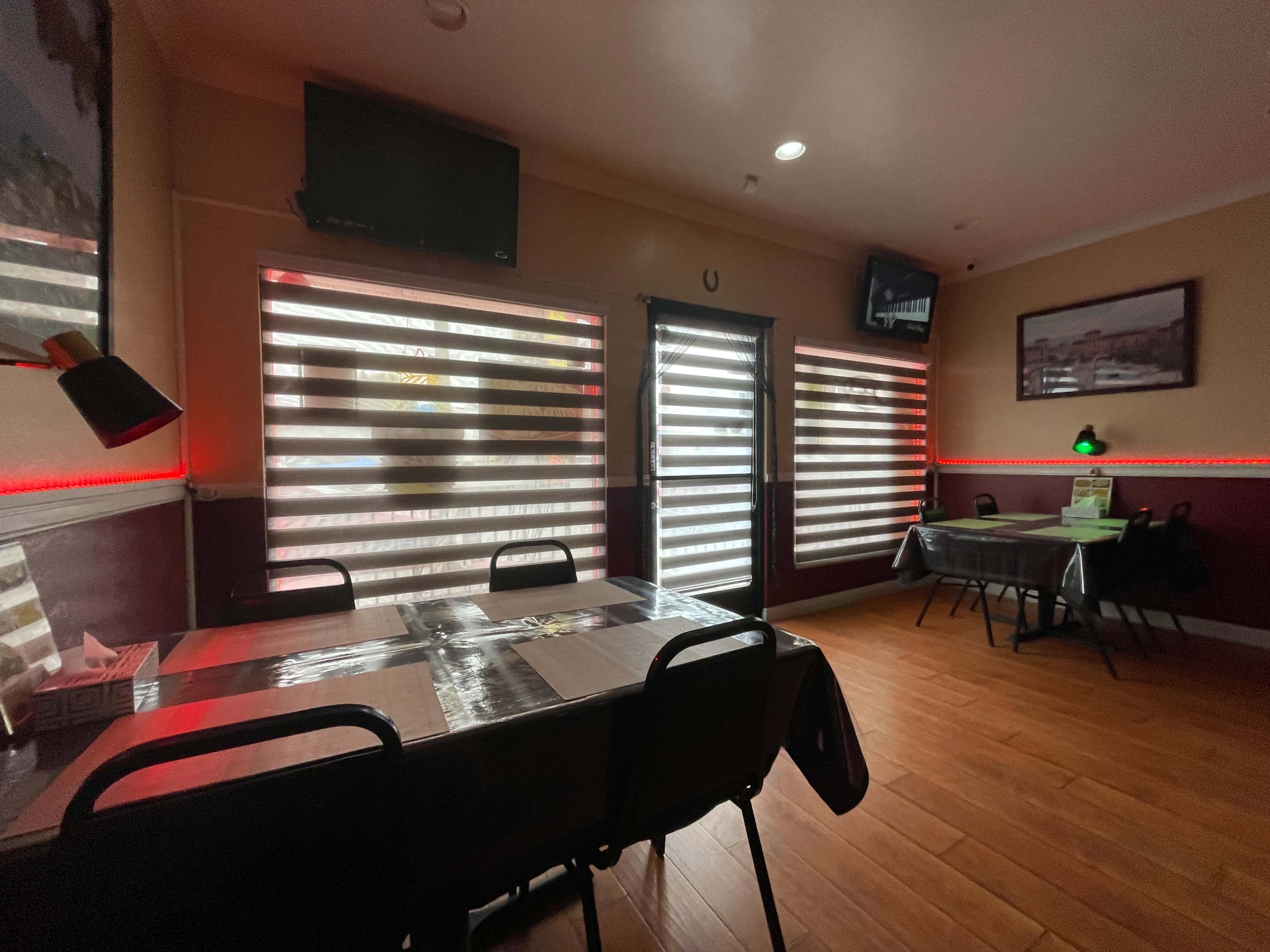 The image shows a restaurant interior with tables and chairs, featuring large windows covered by horizontal blinds and illuminated by dim lighting.