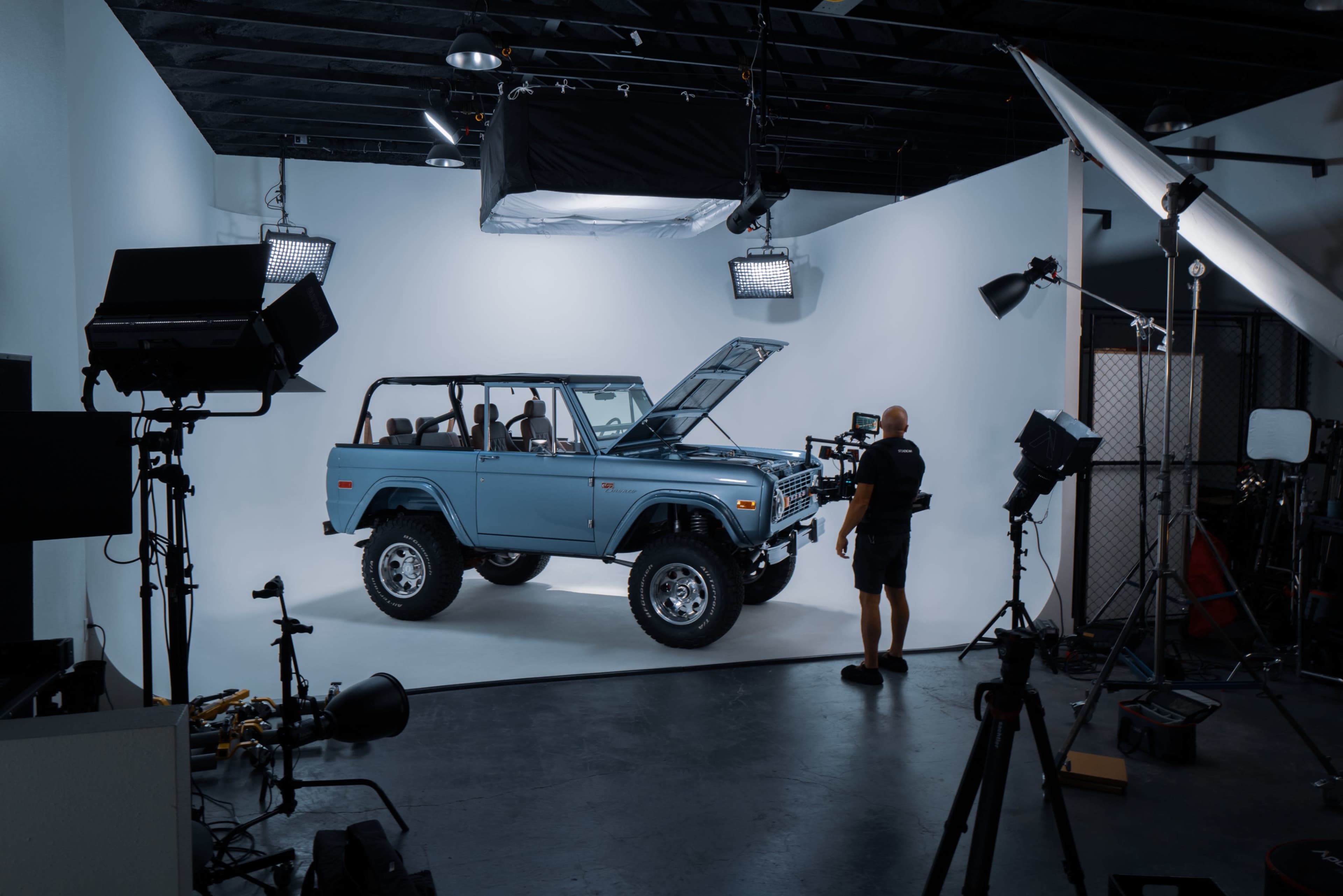 A blue vintage SUV is displayed on a light gray backdrop in a photography studio, with studio lights and equipment around it.