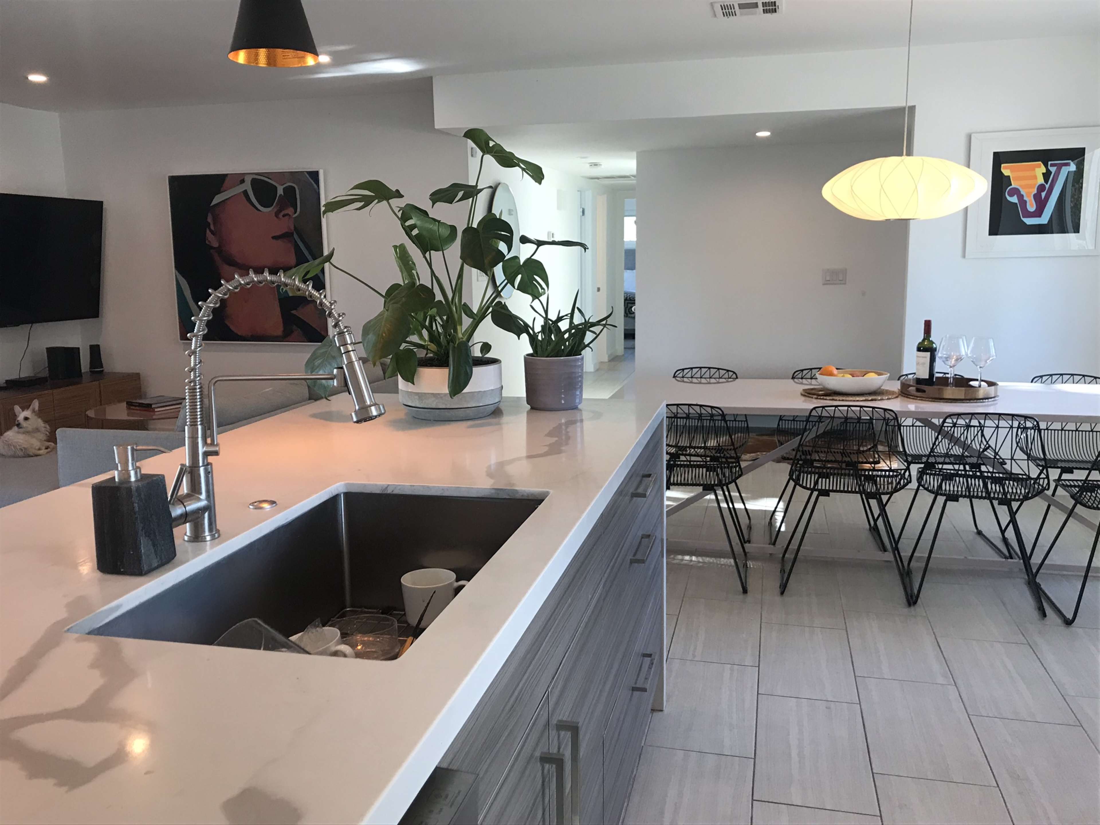 A modern kitchen features a large island with a sink and potted plants, adjacent to a dining area with wire chairs and a bottle of wine on the table.
