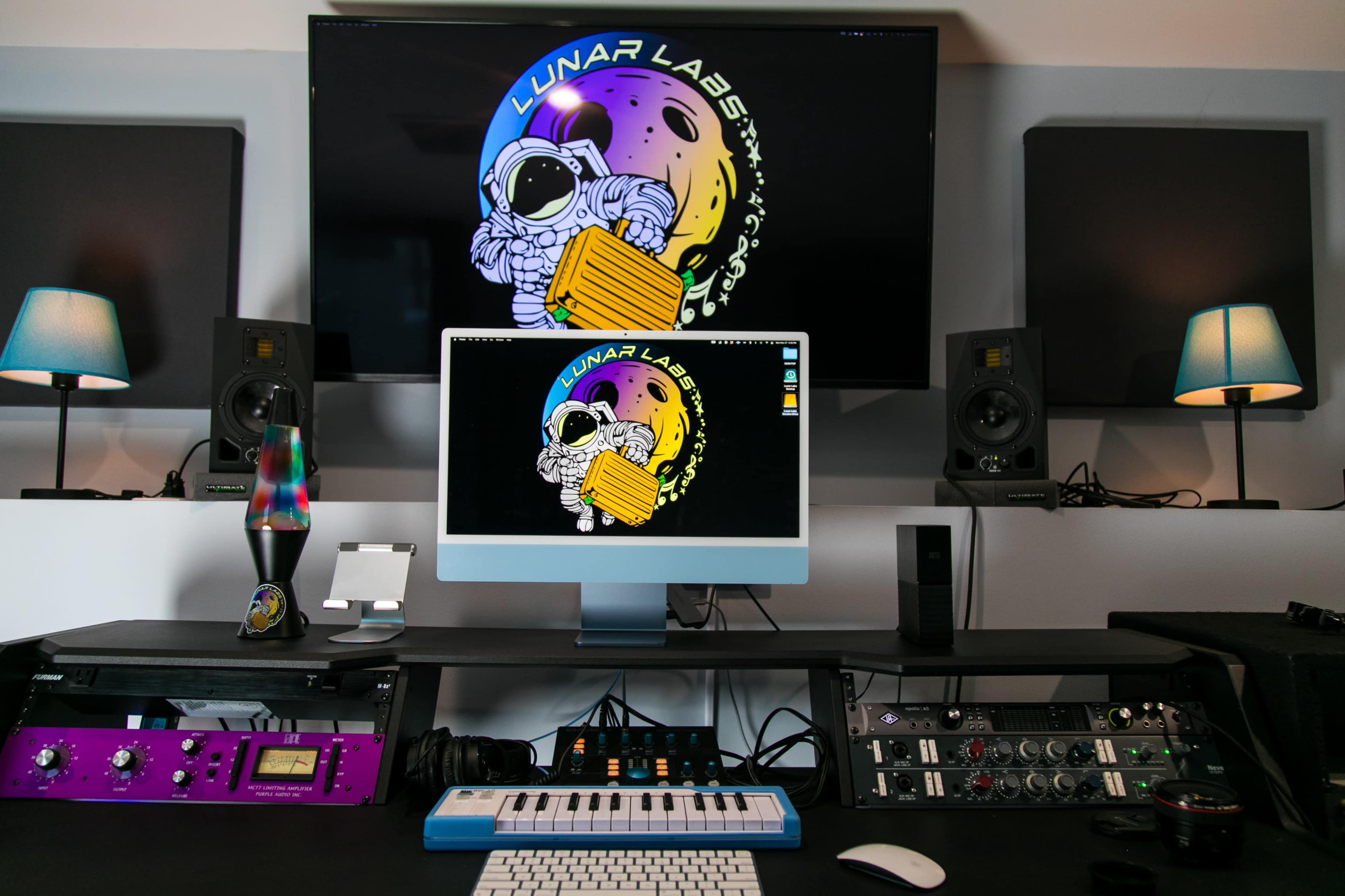 A music studio setup features a computer monitor displaying the "Lunar Labs" logo, surrounded by audio equipment, speakers, and colorful lighting.