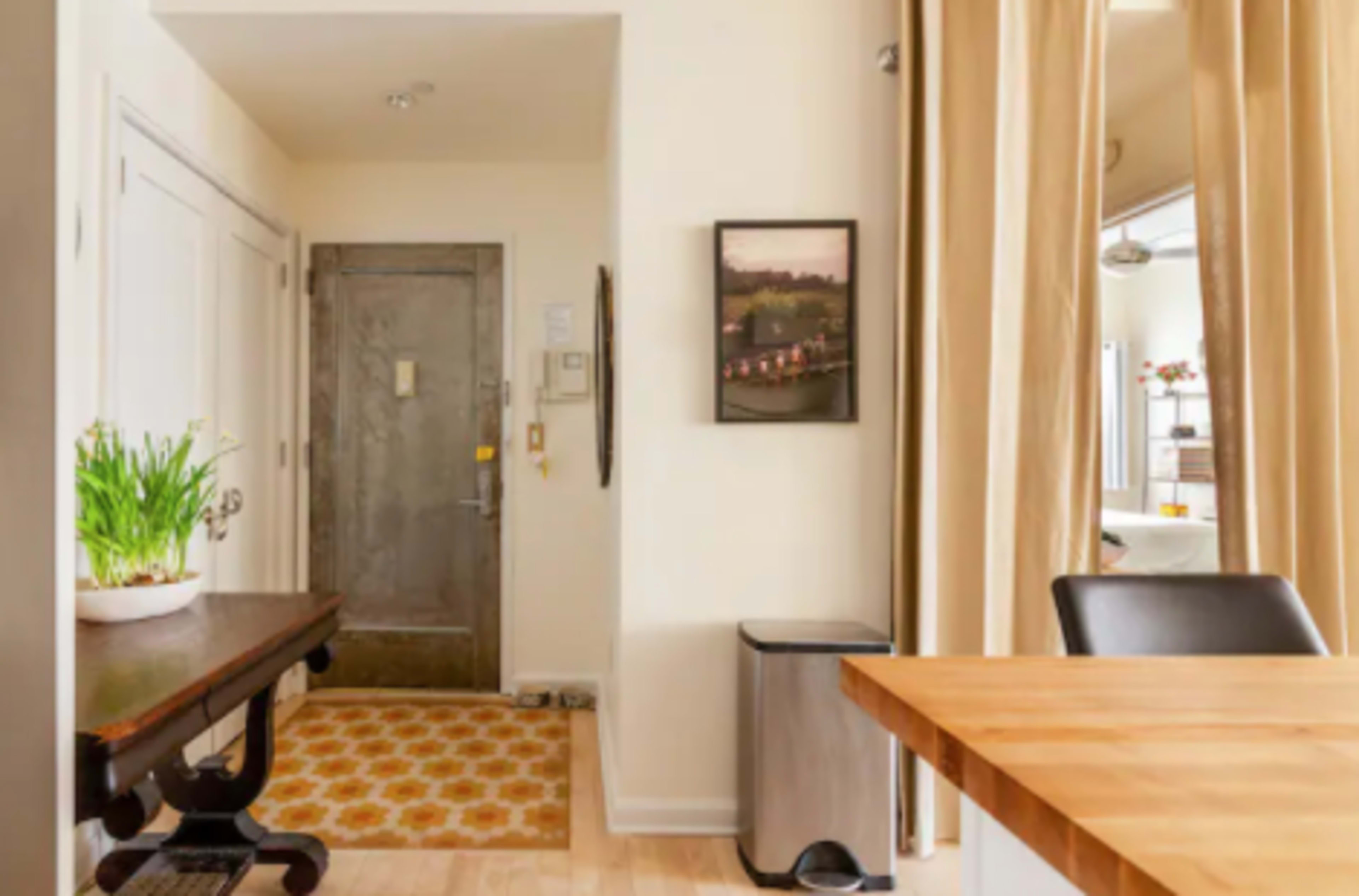 A hallway with a front door, a table on the left, a decorative plant, and a trash can near the wall.