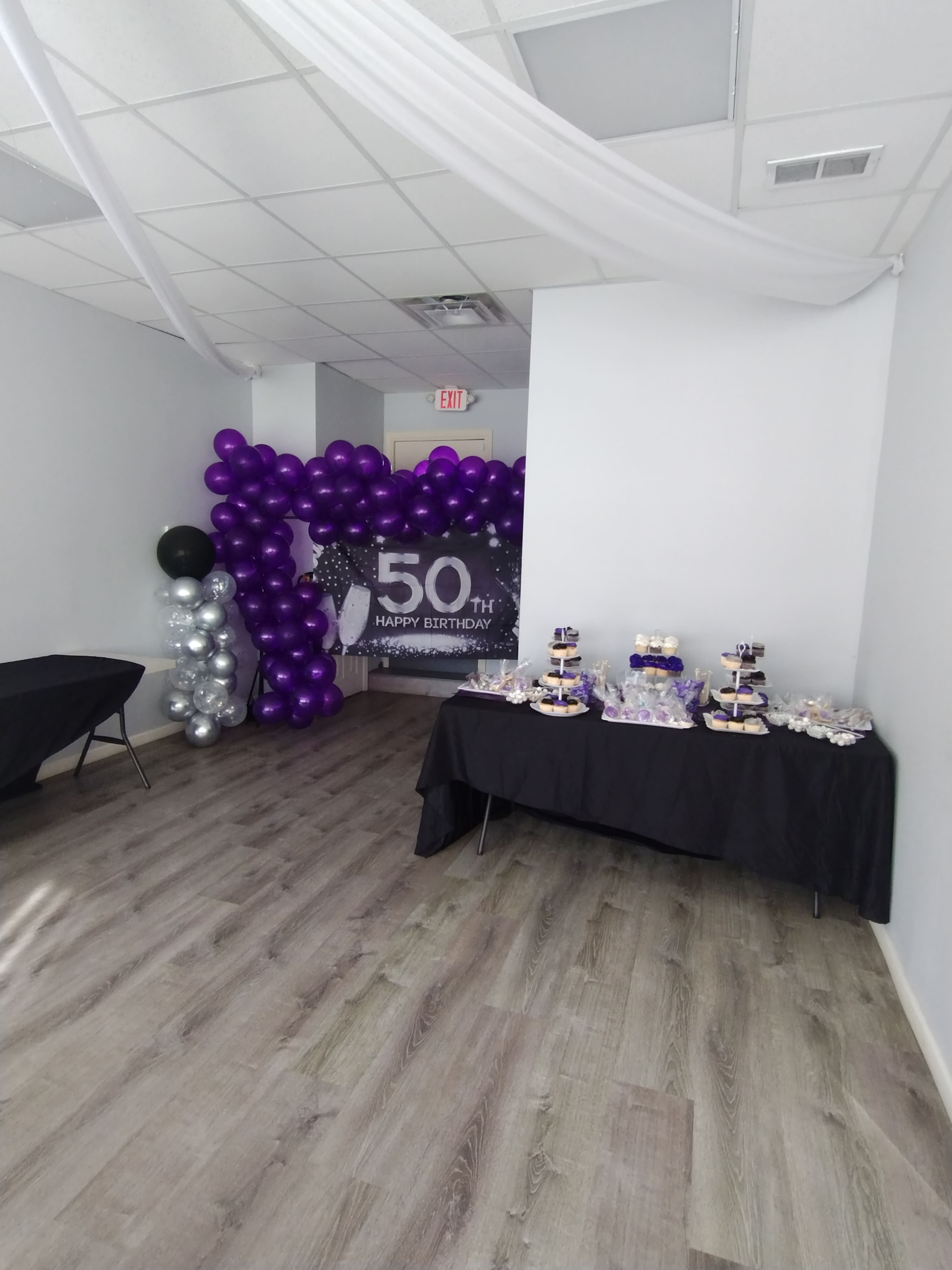 A decorated room for a 50th birthday celebration, featuring a purple balloon arch and dessert tables with a variety of treats.