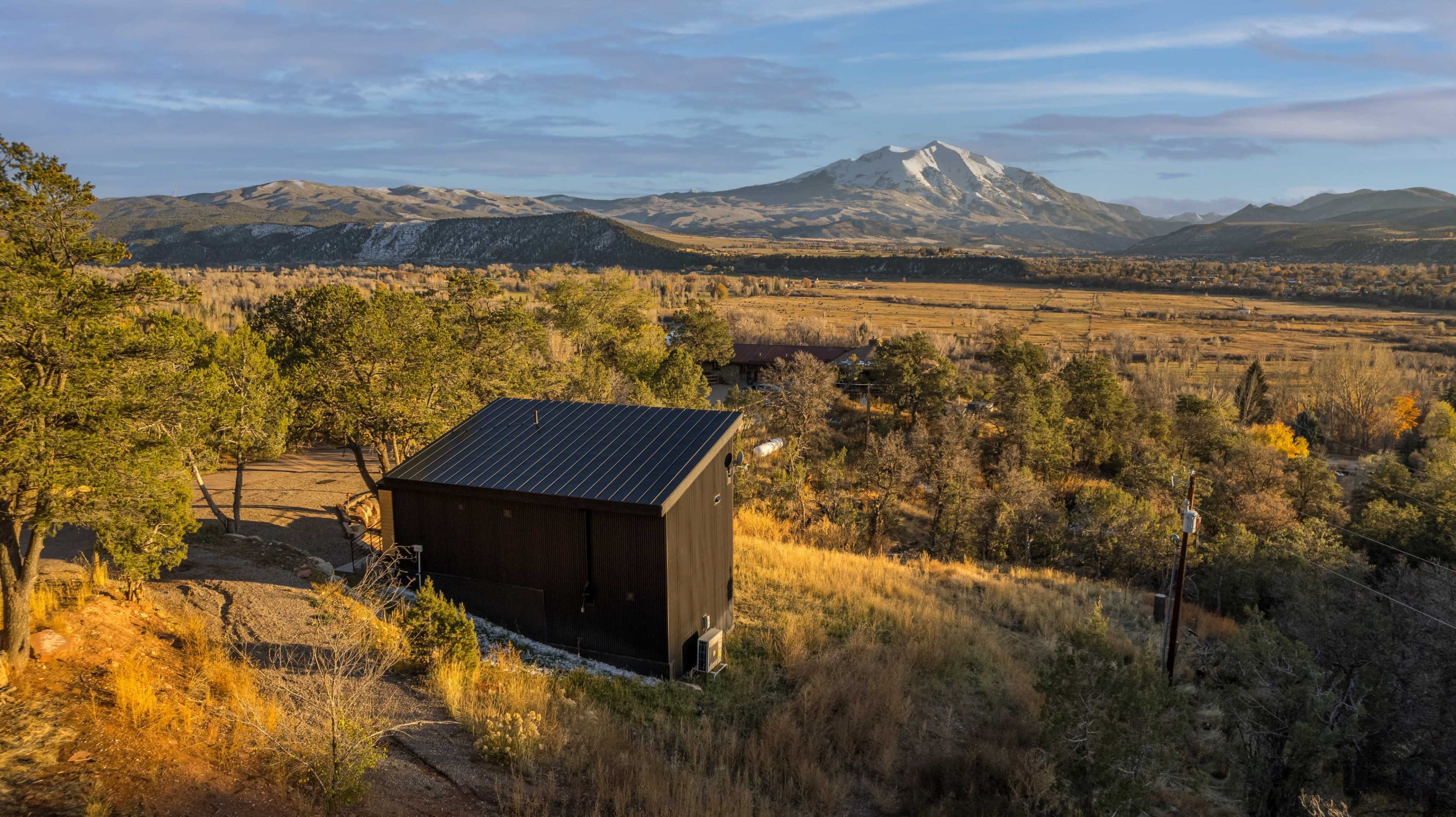 A modern black cabin sits on a hillside overlooking a valley with mountains in the background under a blue sky.