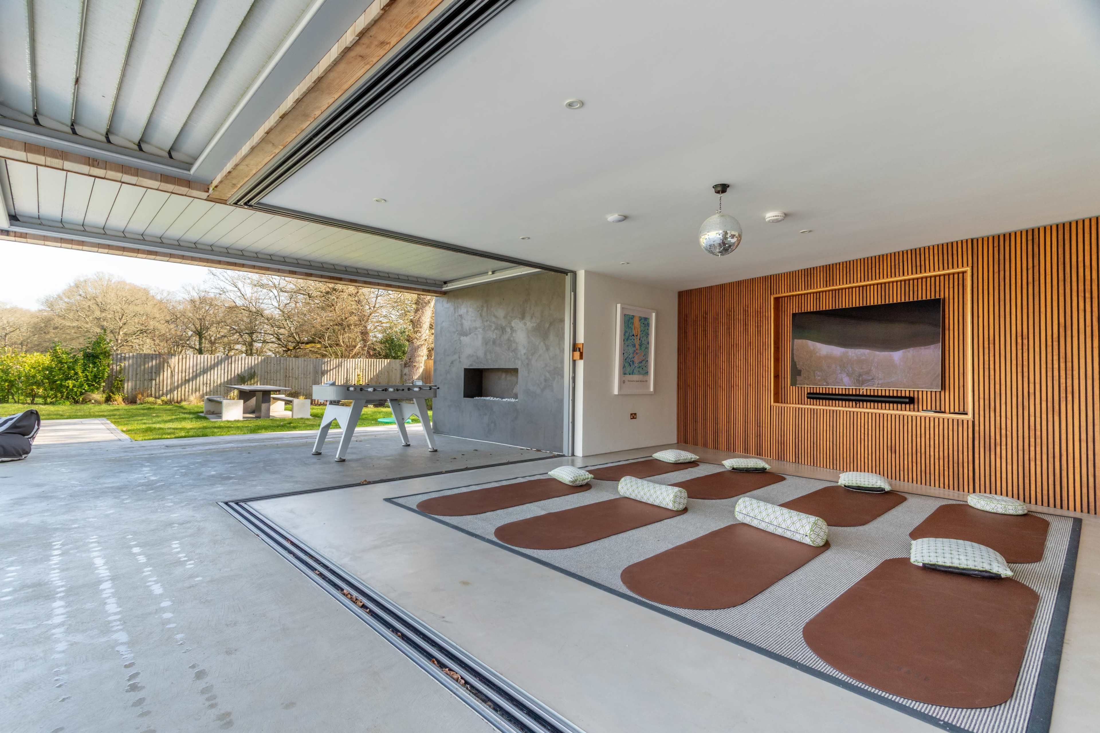 A spacious indoor area features a large opening to an outdoor space, with several cushioned seating pads arranged on a smooth concrete floor and a television mounted on a wooden wall.