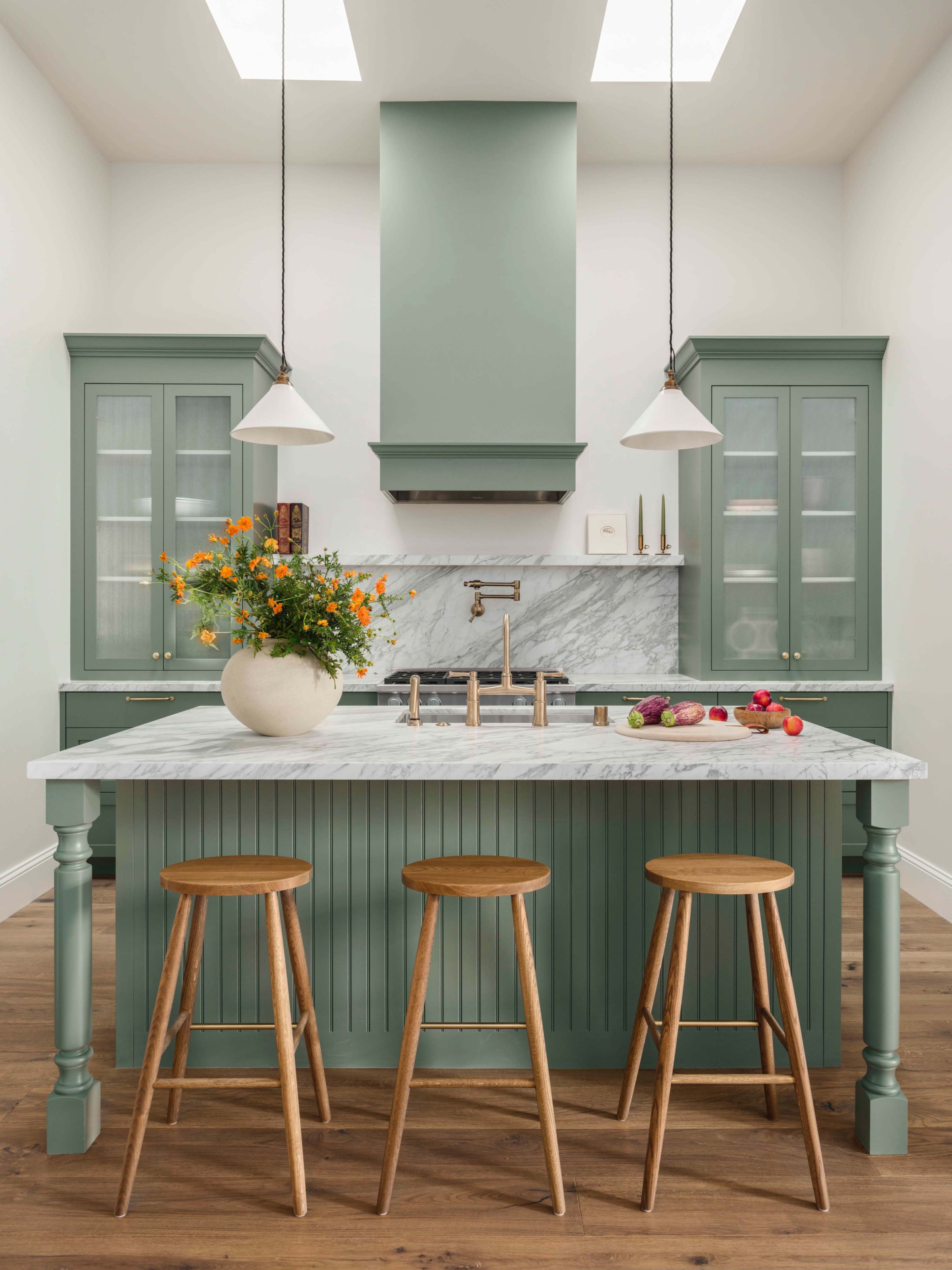 The image shows a modern kitchen with a green island, marble countertop, three wooden stools, and floral arrangements on the island.