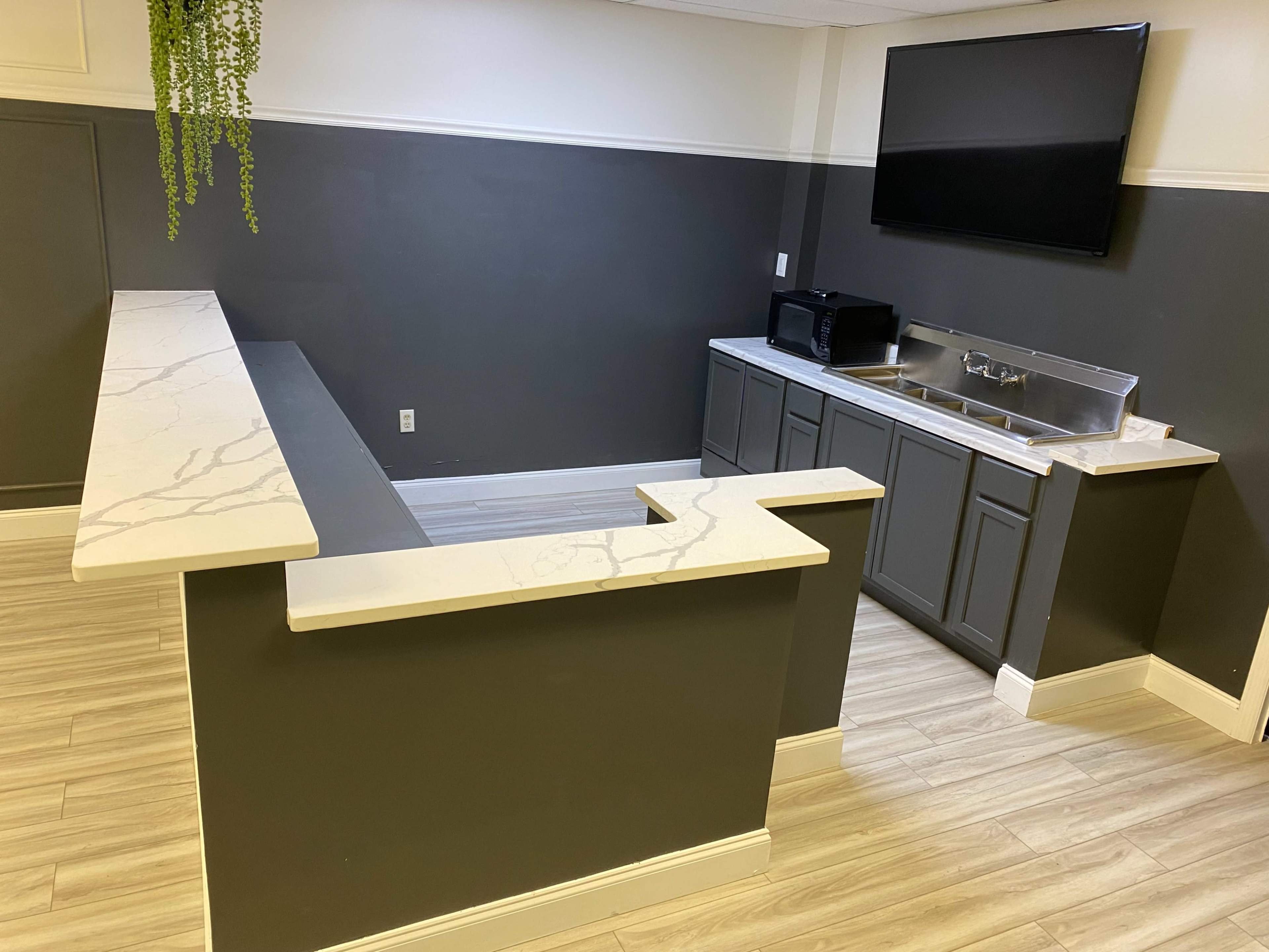 A modern kitchenette area featuring a countertop, stainless steel sink, microwave, and a large wall-mounted TV.