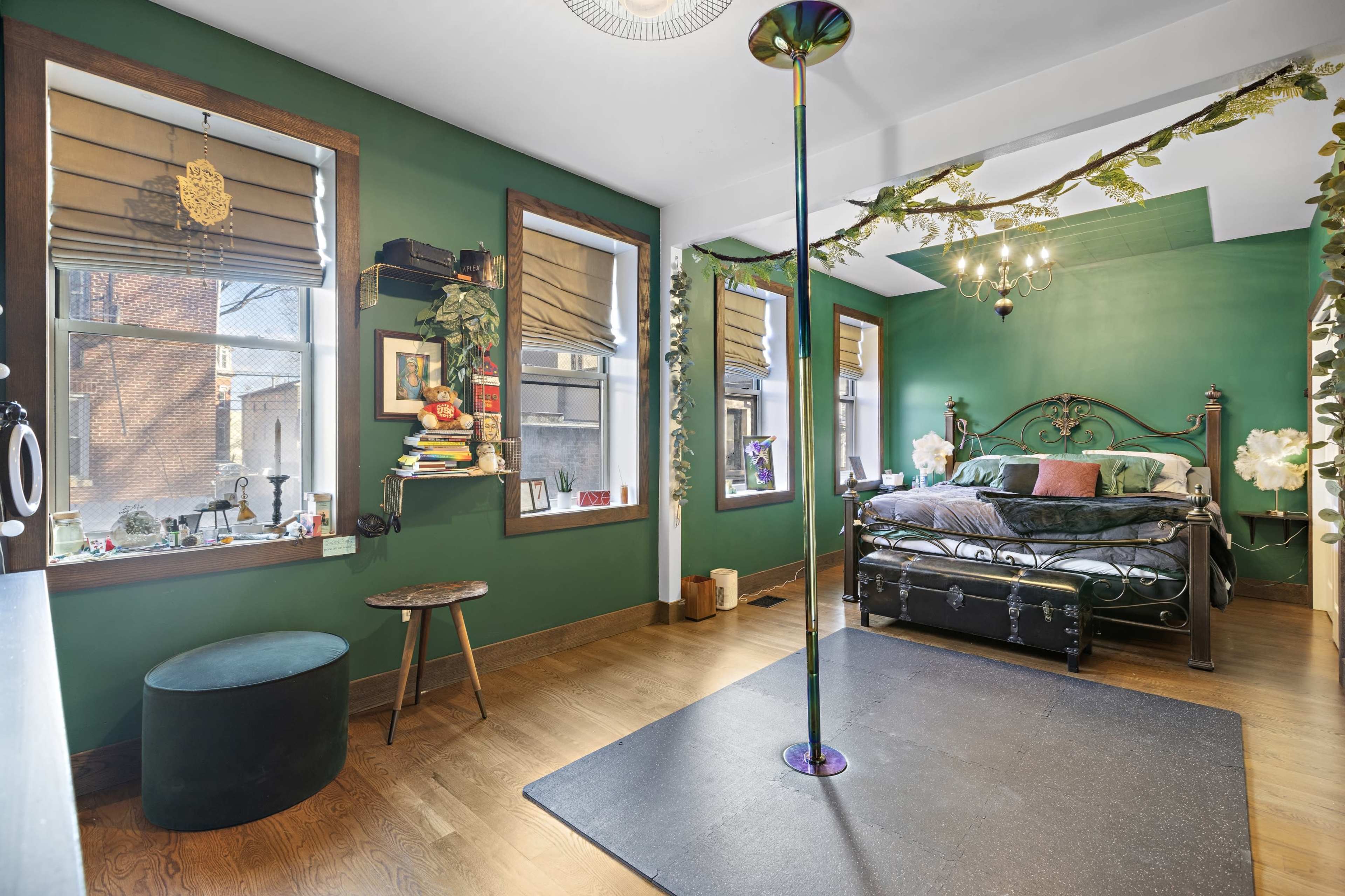 The image shows a dark green bedroom featuring a black metal bed, a standing pole, wooden flooring, and large windows with blinds.