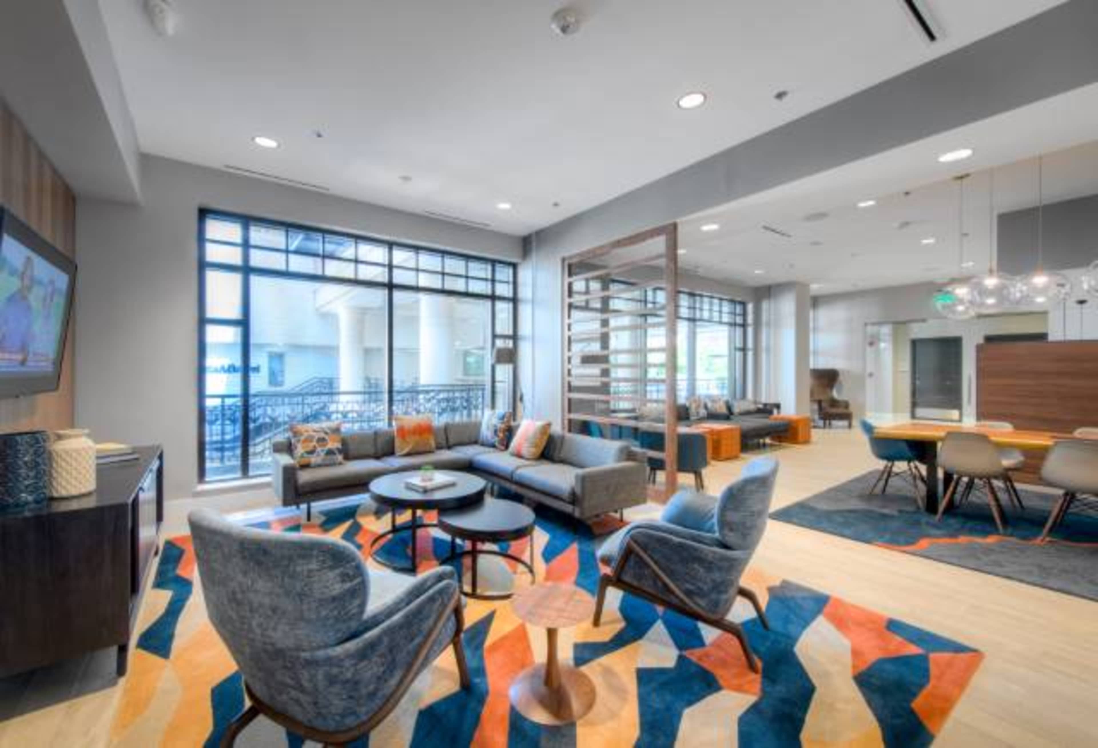 A modern lounge area features comfortable seating, a large rug with a geometric pattern, and a dining space with a round table surrounded by chairs.
