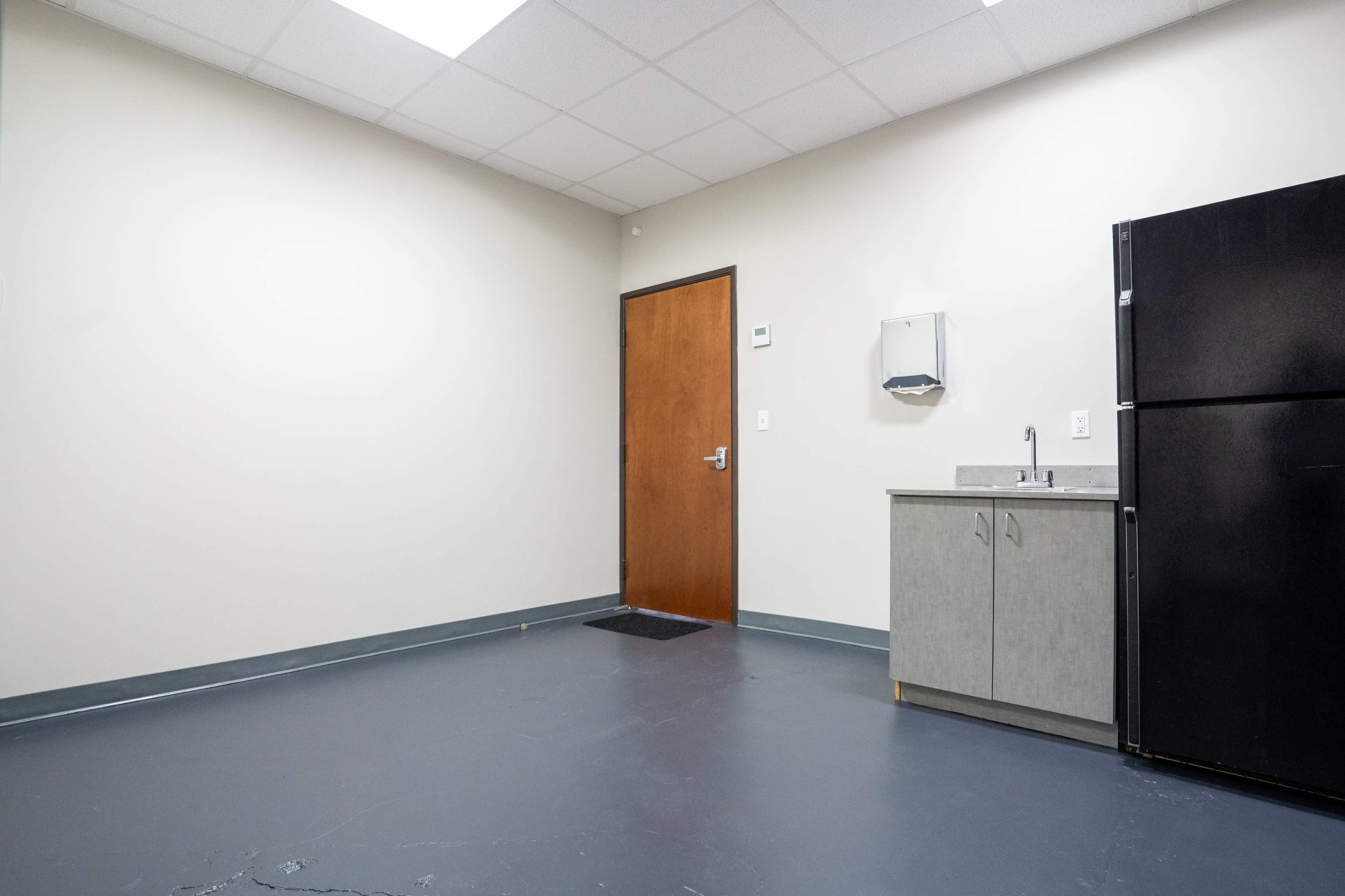 An empty room features a gray kitchenette with a sink and a black refrigerator, along with a door on the adjacent wall.