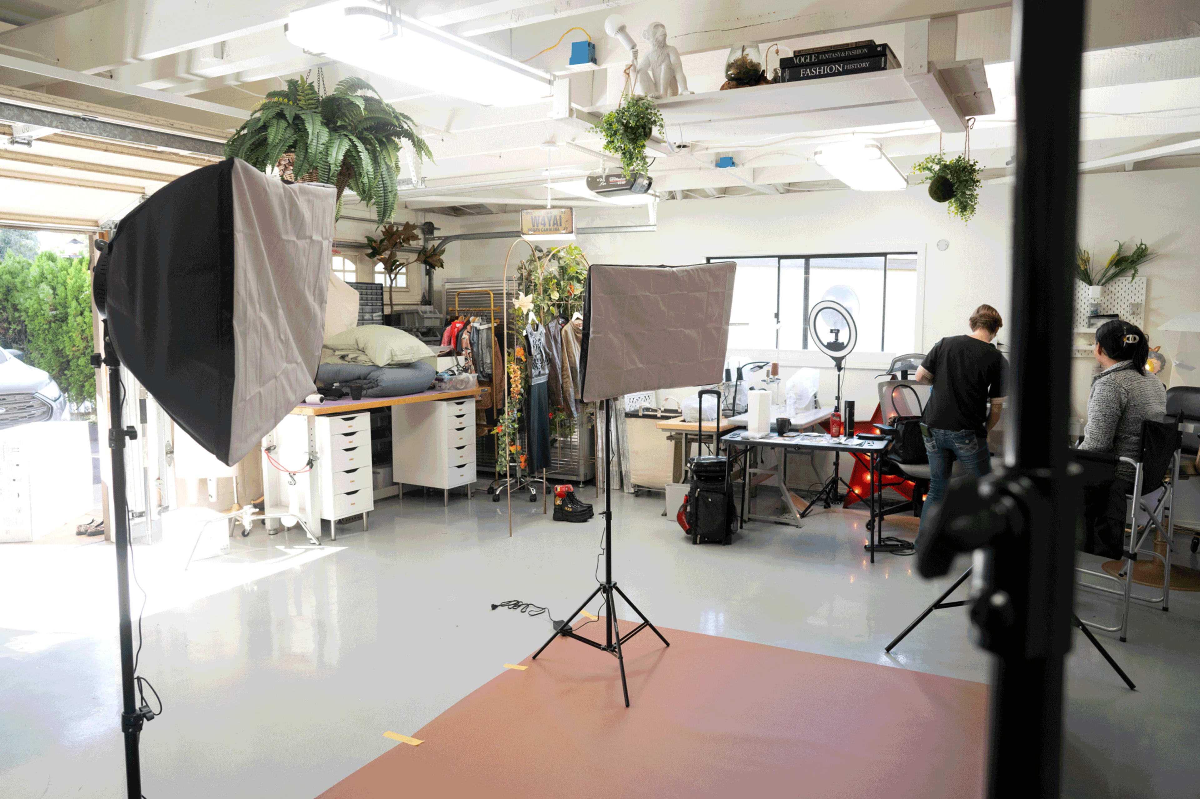 A well-lit studio space with photography equipment set up, including softboxes, and two people working at a table amidst various props and plants.