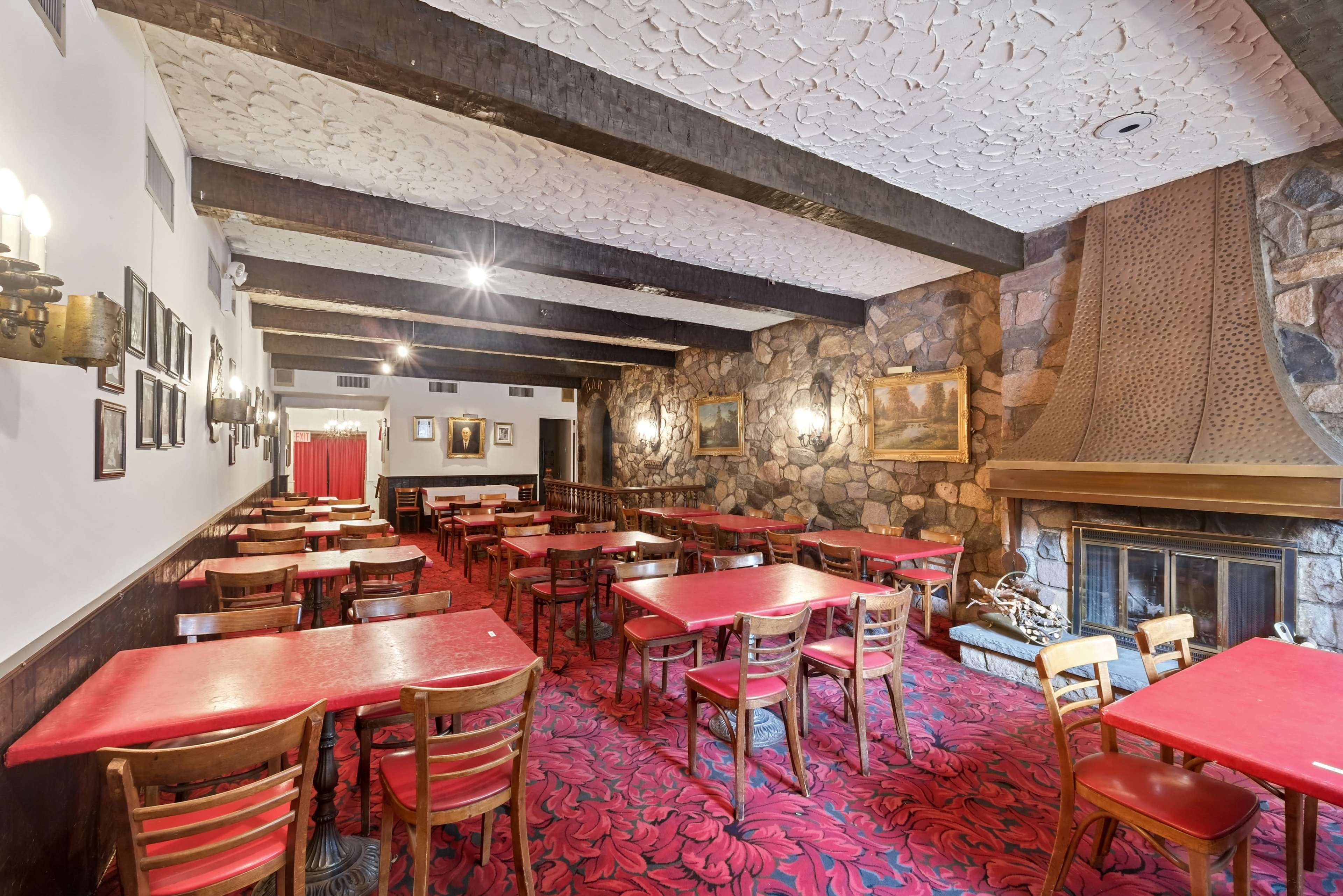 A restaurant interior featuring red tables and chairs, a stone fireplace, and textured walls with wooden beams overhead.