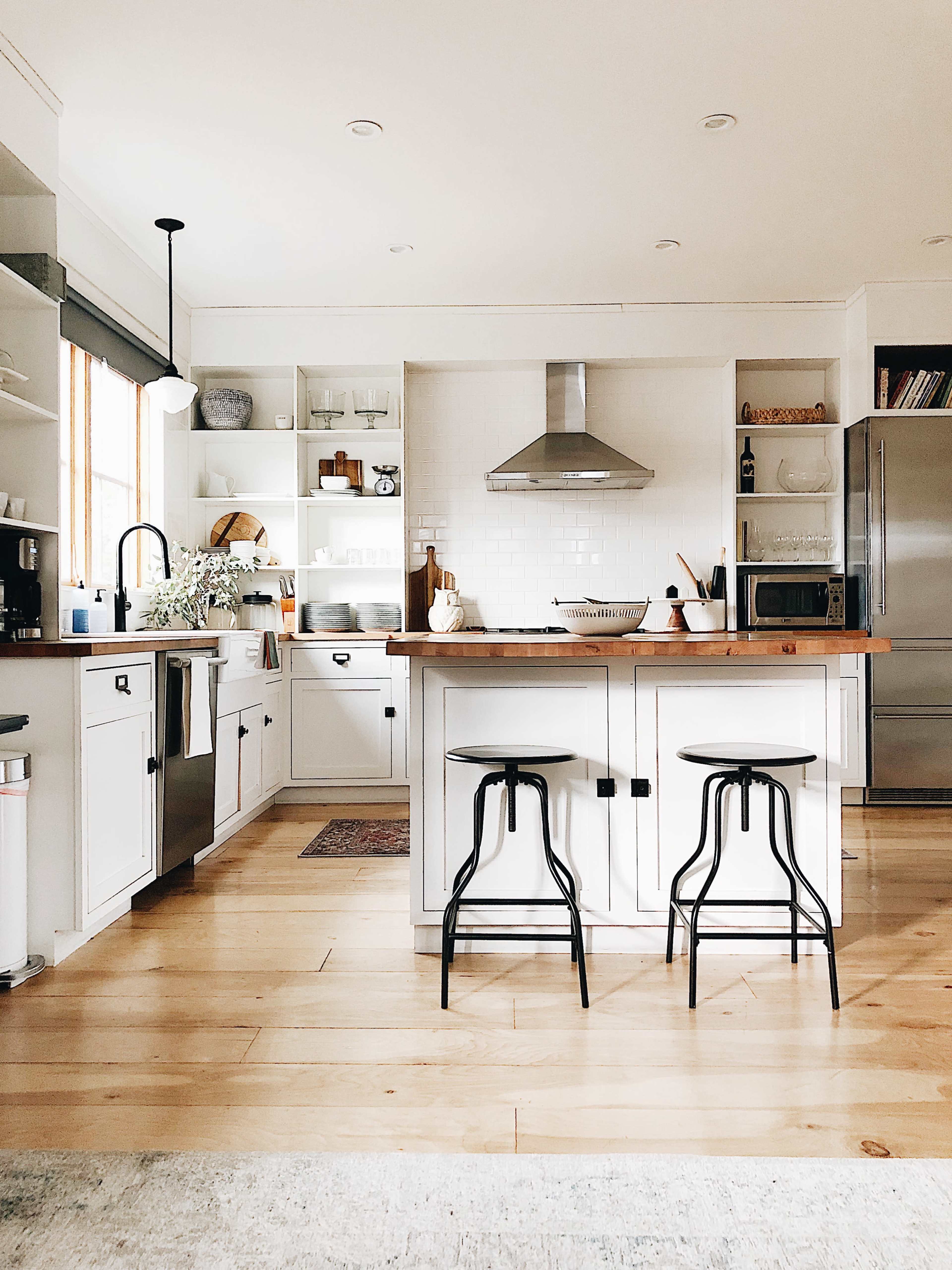 A modern kitchen features white cabinetry, a central island with wooden countertops, sleek stools, and stainless steel appliances.