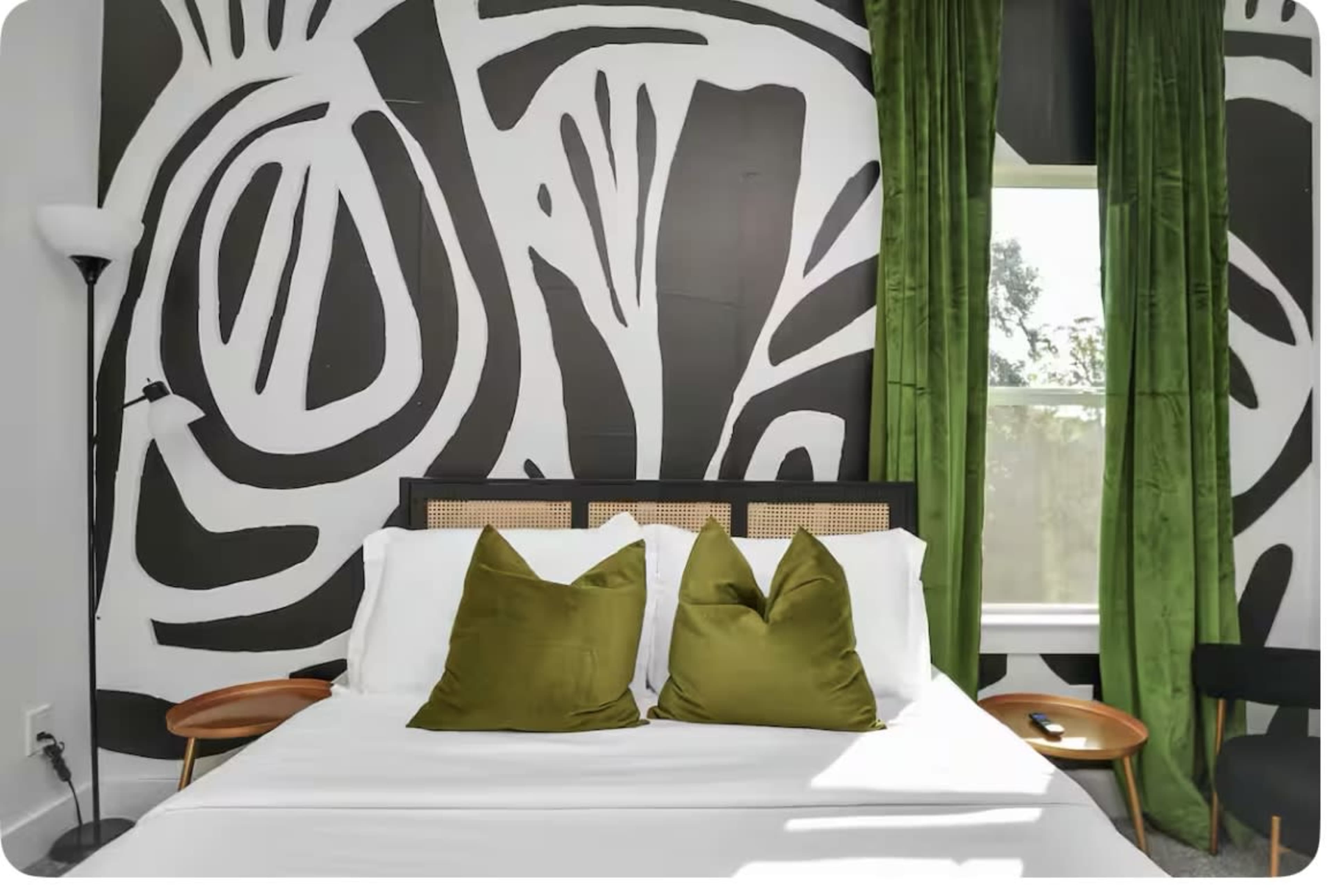 The image shows a bedroom with a bed featuring two green accent pillows, a black and white patterned wall, and green curtains.