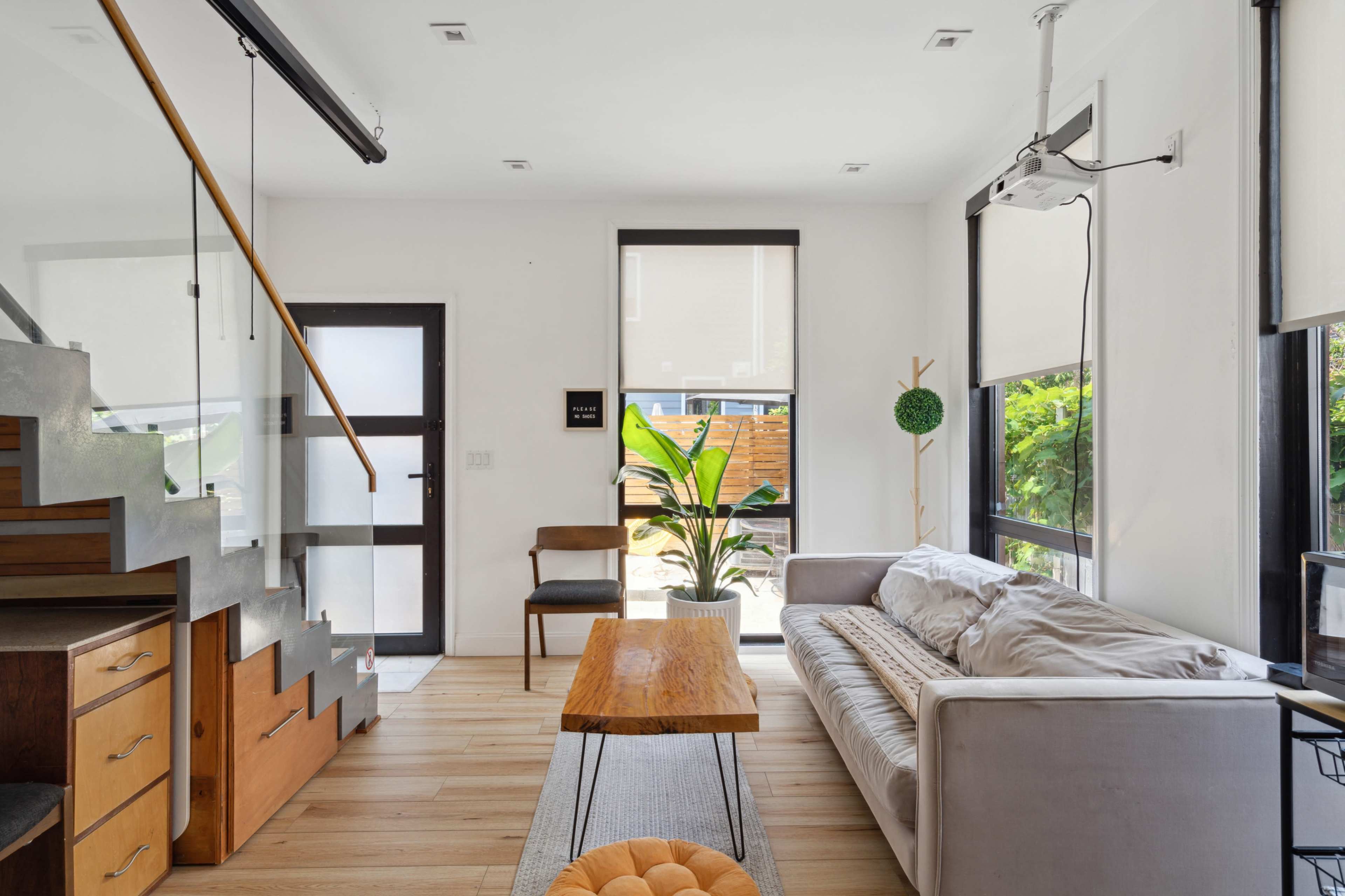 The image depicts a modern living room featuring a staircase, a coffee table, a sofa, and a potted plant, with large windows allowing natural light to enter.