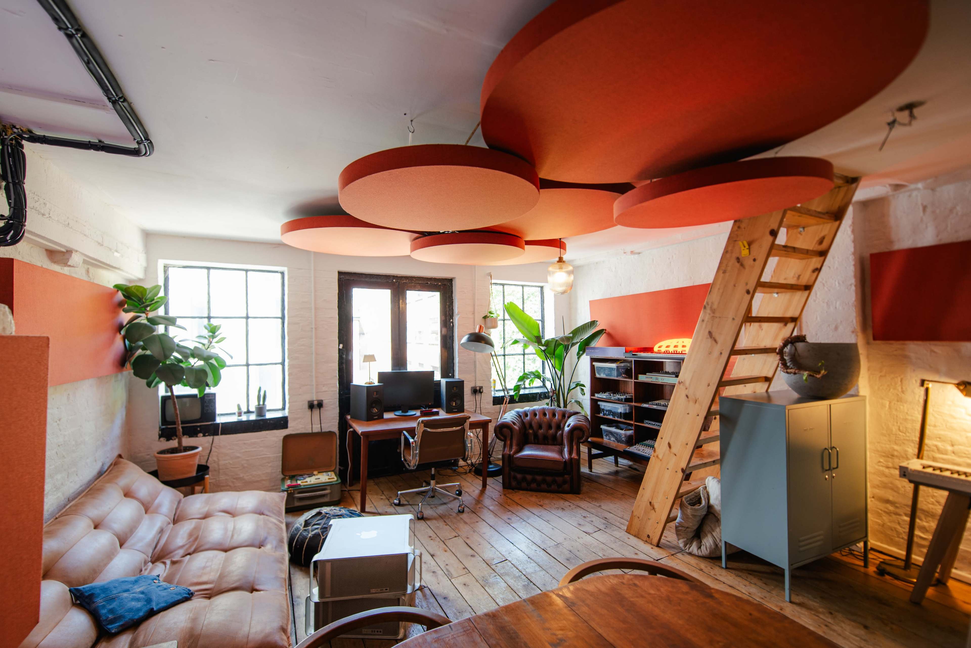 The image shows a cozy, modern workspace with a leather couch, a staircase, several green plants, and acoustic panels hanging from the ceiling.