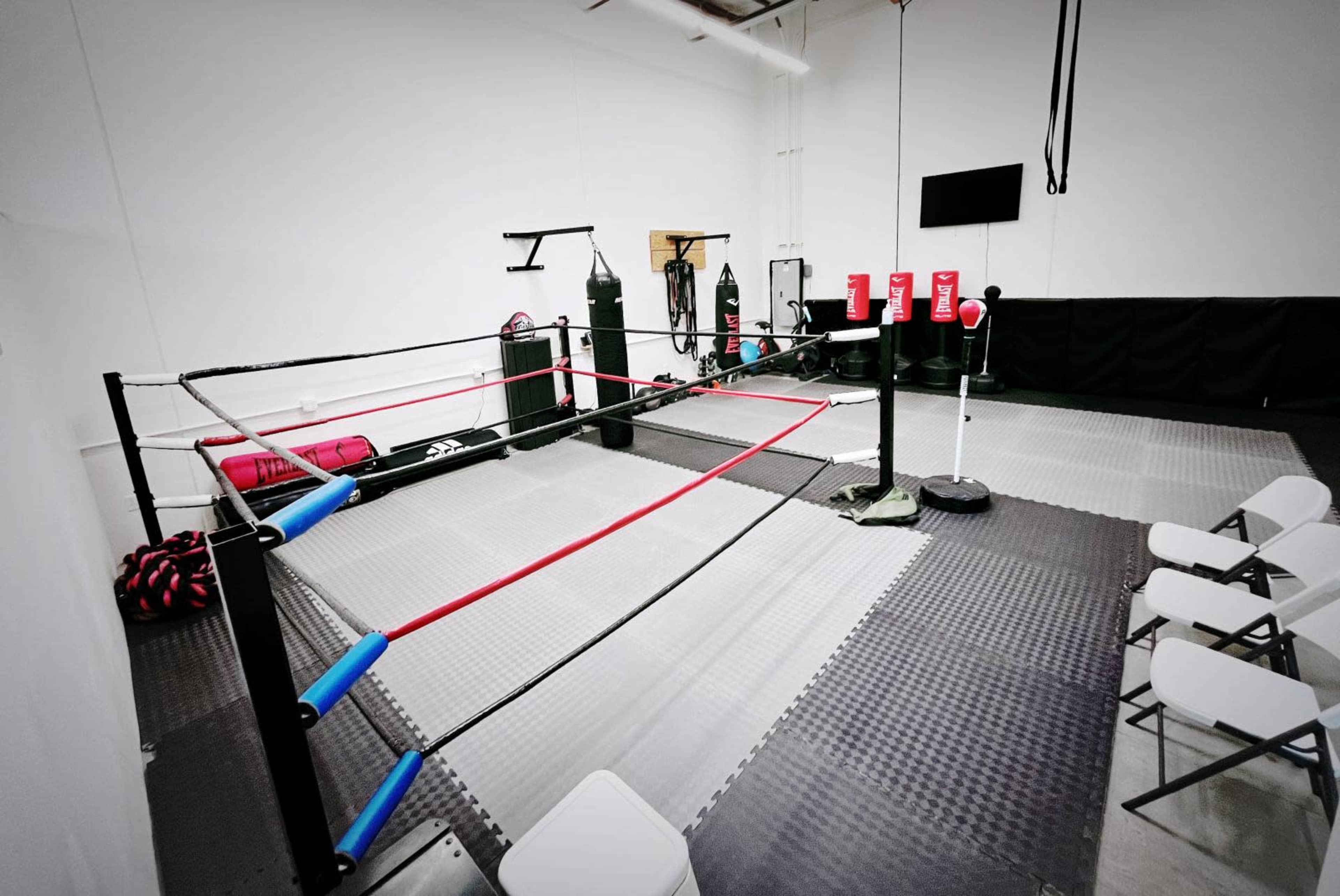 The image shows a boxing training area featuring a ring, punching bags, and several chairs arranged nearby.
