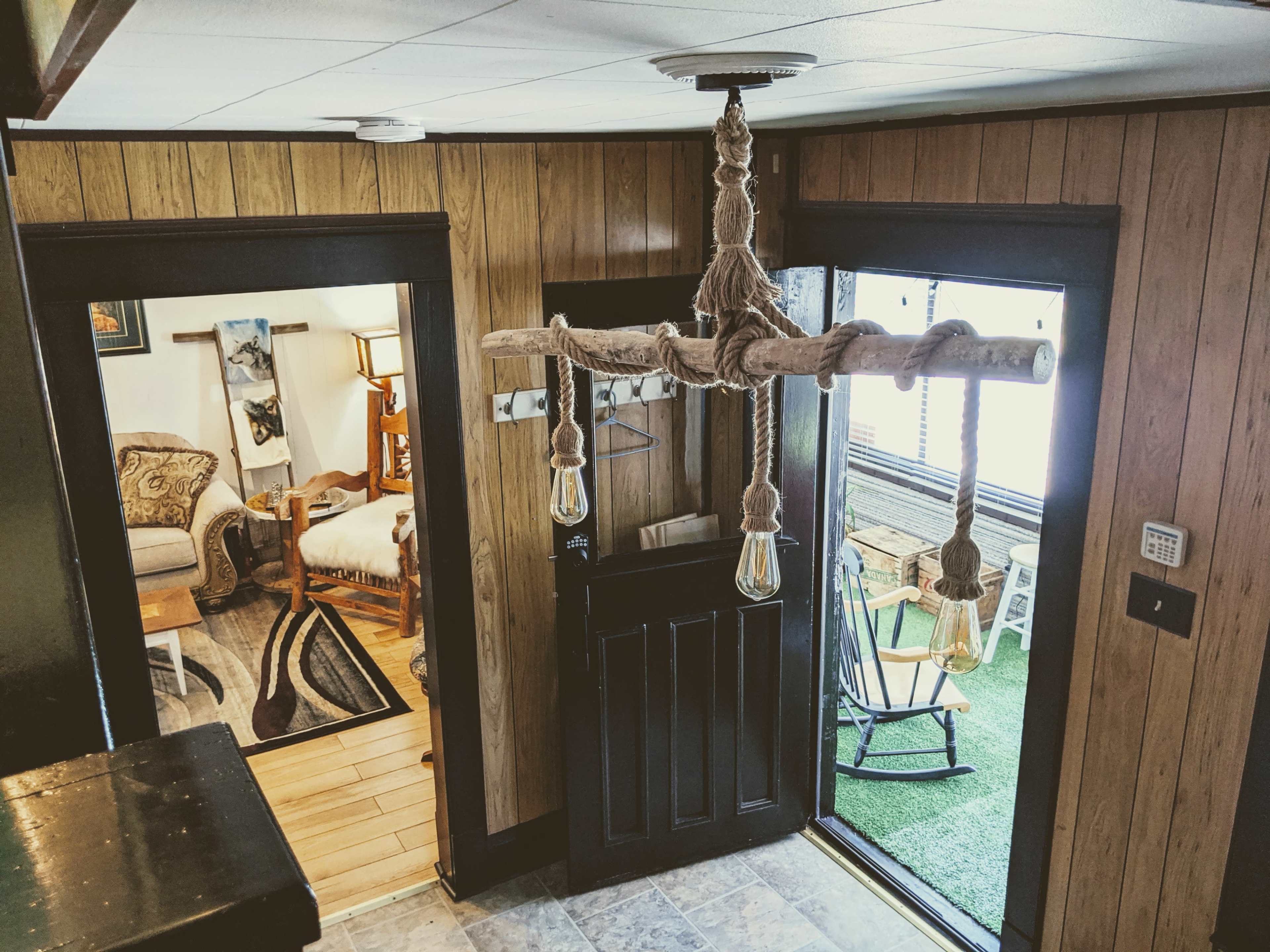 A hallway with a wooden chandelier hanging from the ceiling, leading to two doorways: one into a cozy room with a bed and armchair, and another opening to a small outdoor area with a rocking chair.