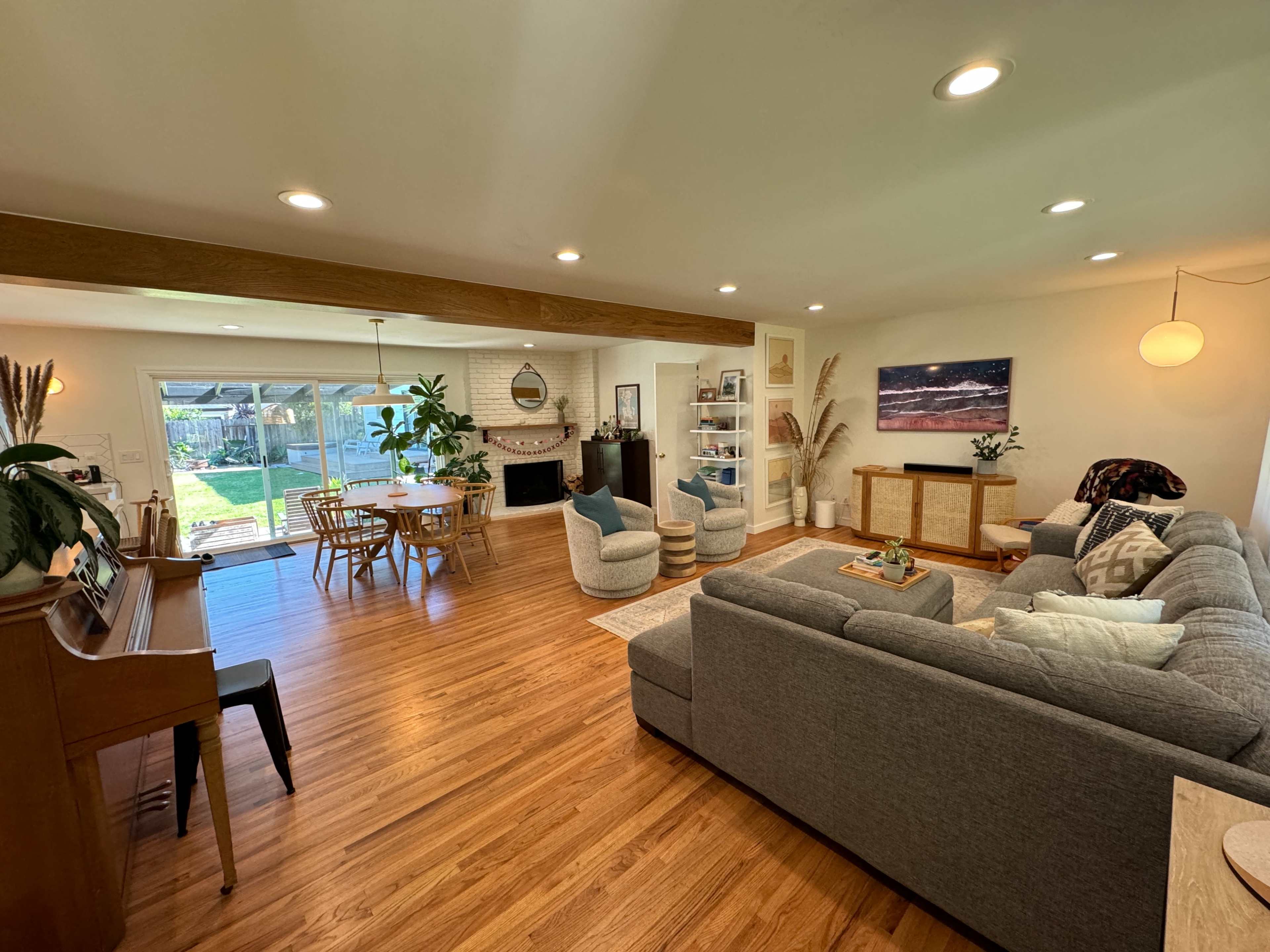 A modern living room with a sectional sofa, piano, and a dining area that opens to a backyard through large glass doors.