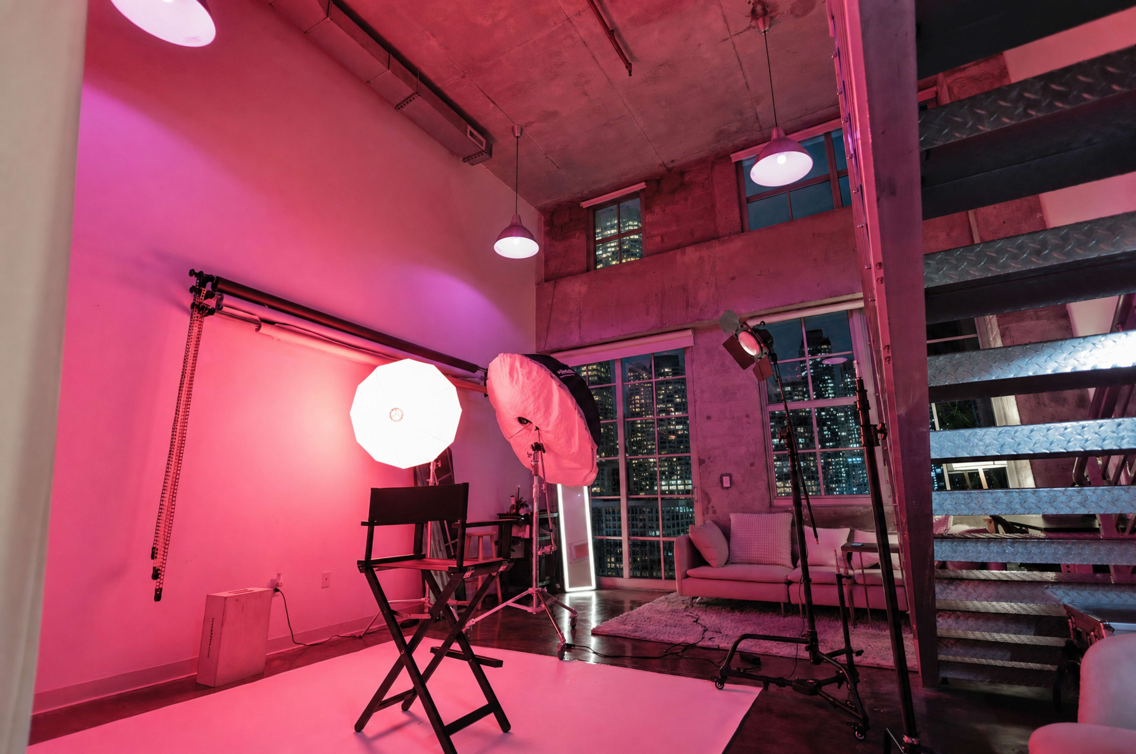 The image shows a modern photography studio setup with various lighting equipment, a director's chair, and a large window revealing a city skyline.